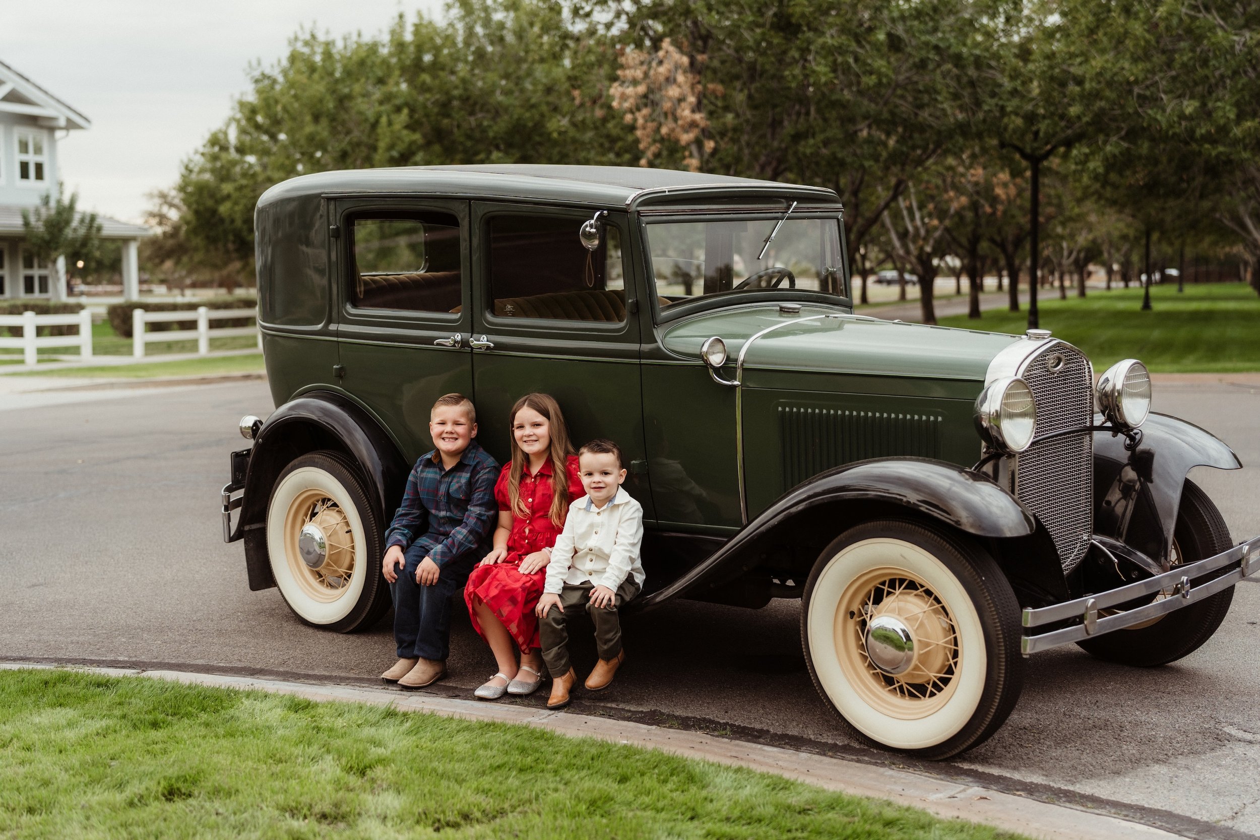 Gabby Lane Classics family photos of three kids sitting in front of a vintage 1931 Ford Four Door ("Green") car on a suburban street with trees and houses in the background.