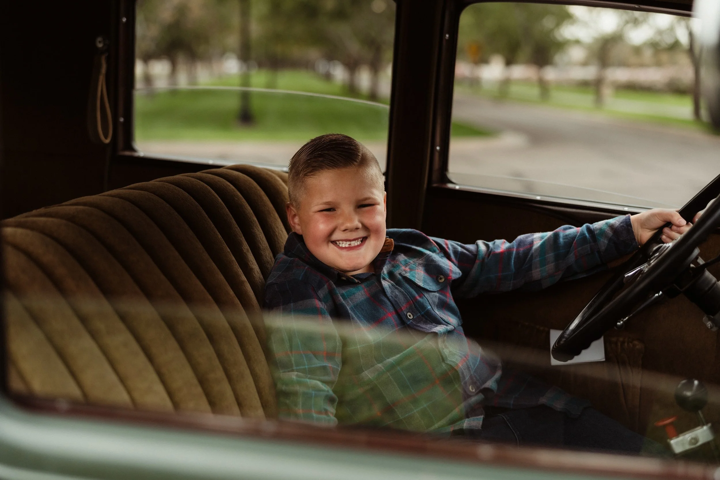 Gabby lane Classics family photo of a young boy sitting in the driver's seat of a vintage 1931 Ford Four Door ("Green") vehicle, holding the steering wheel inside a brown interior, with a park and street view visible through the windows.