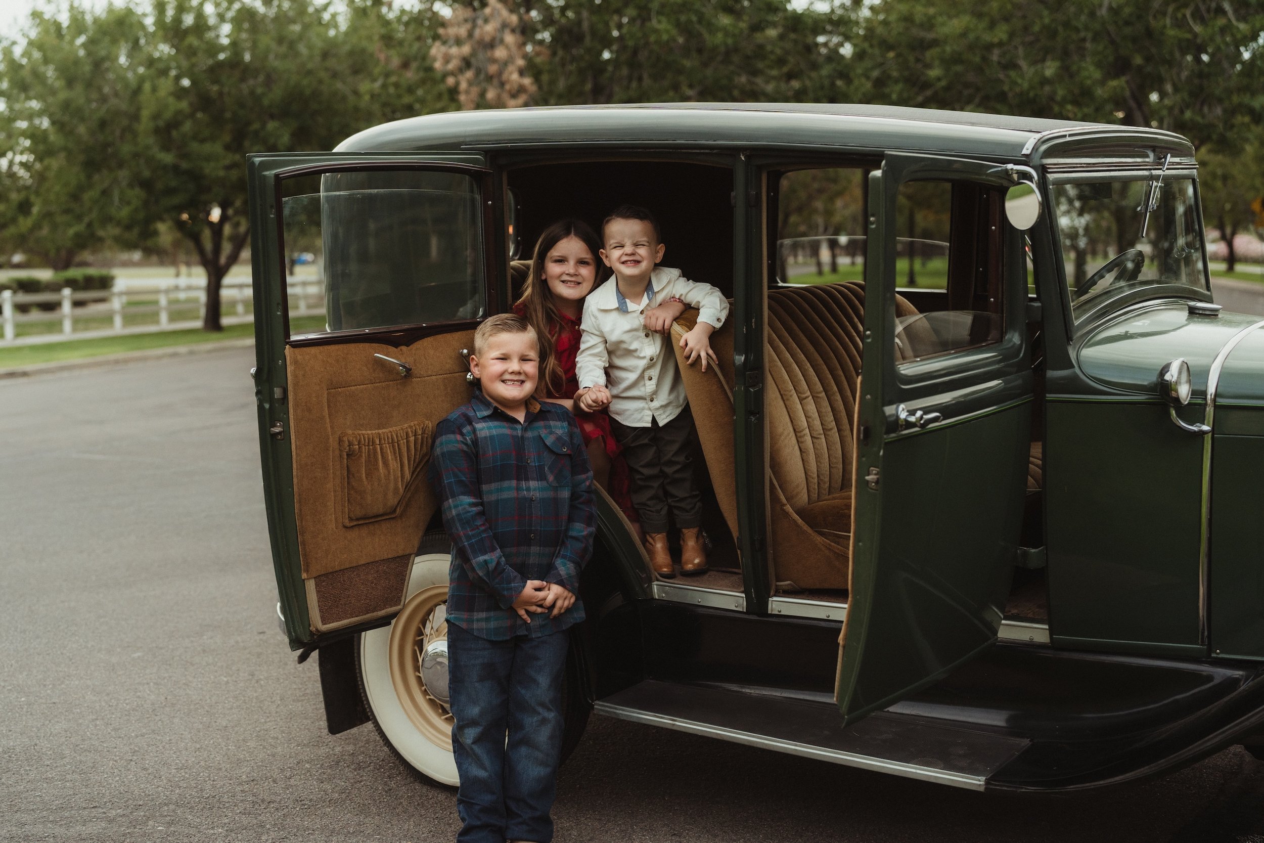 Gabby Lane Classics family photo of three kids smiling and posing in an vintage 1931 Ford Four Door ("Green") car with brown interior, on a paved road in an outdoor park with trees and a white fence in the background.