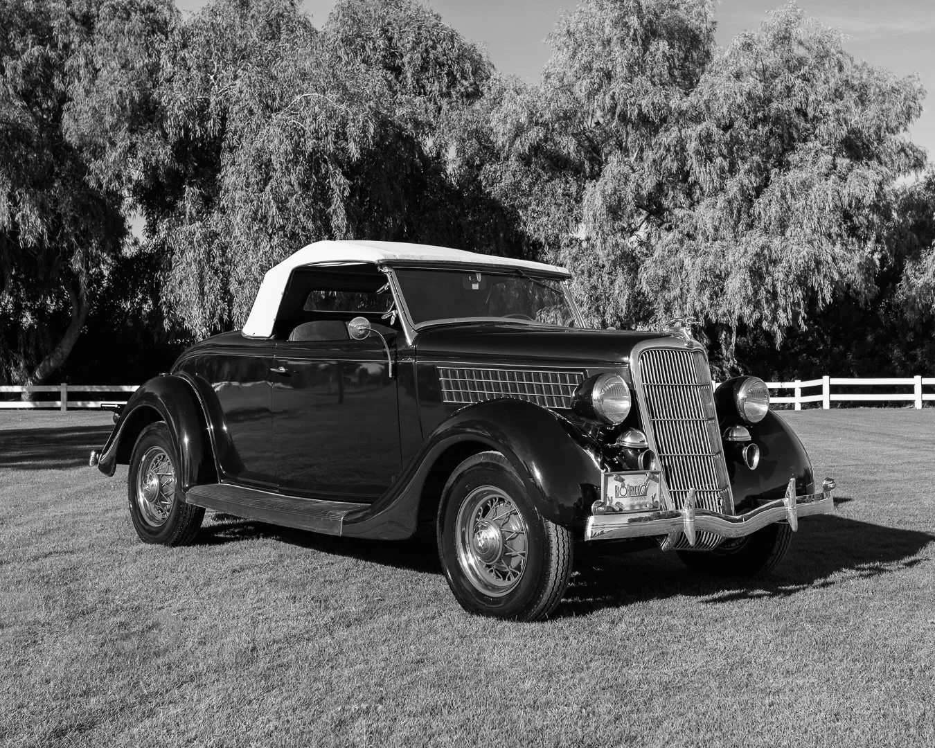A vintage black 1935 Ford Roadster car with a white soft top parked on grass, with large trees and a white fence in the background. Classic car experiences by Gabby Lane Classics.