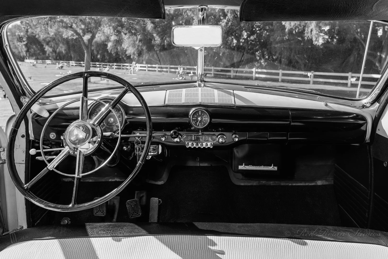 Black and white photo of a vintage 1950 Ford Crestliner car interior showing a large steering wheel, dashboard with gauges, and a view through the windshield of trees and a fence.