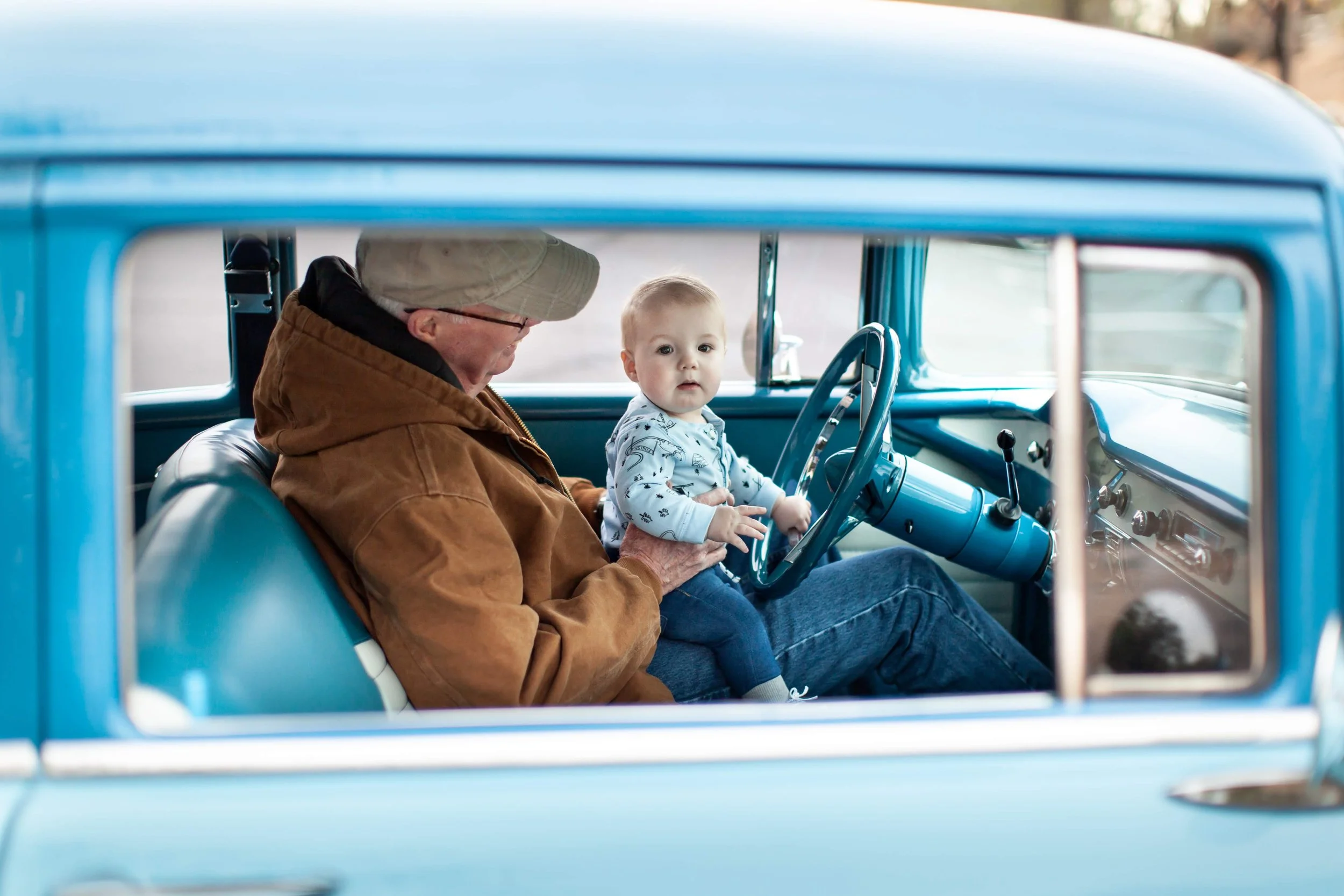 A Gabby Lane Classics photoshoot featuring an elderly man and a young child sit inside a vintage blue car. The child is sitting on the man's lap and holding the steering wheel, looking at the camera.