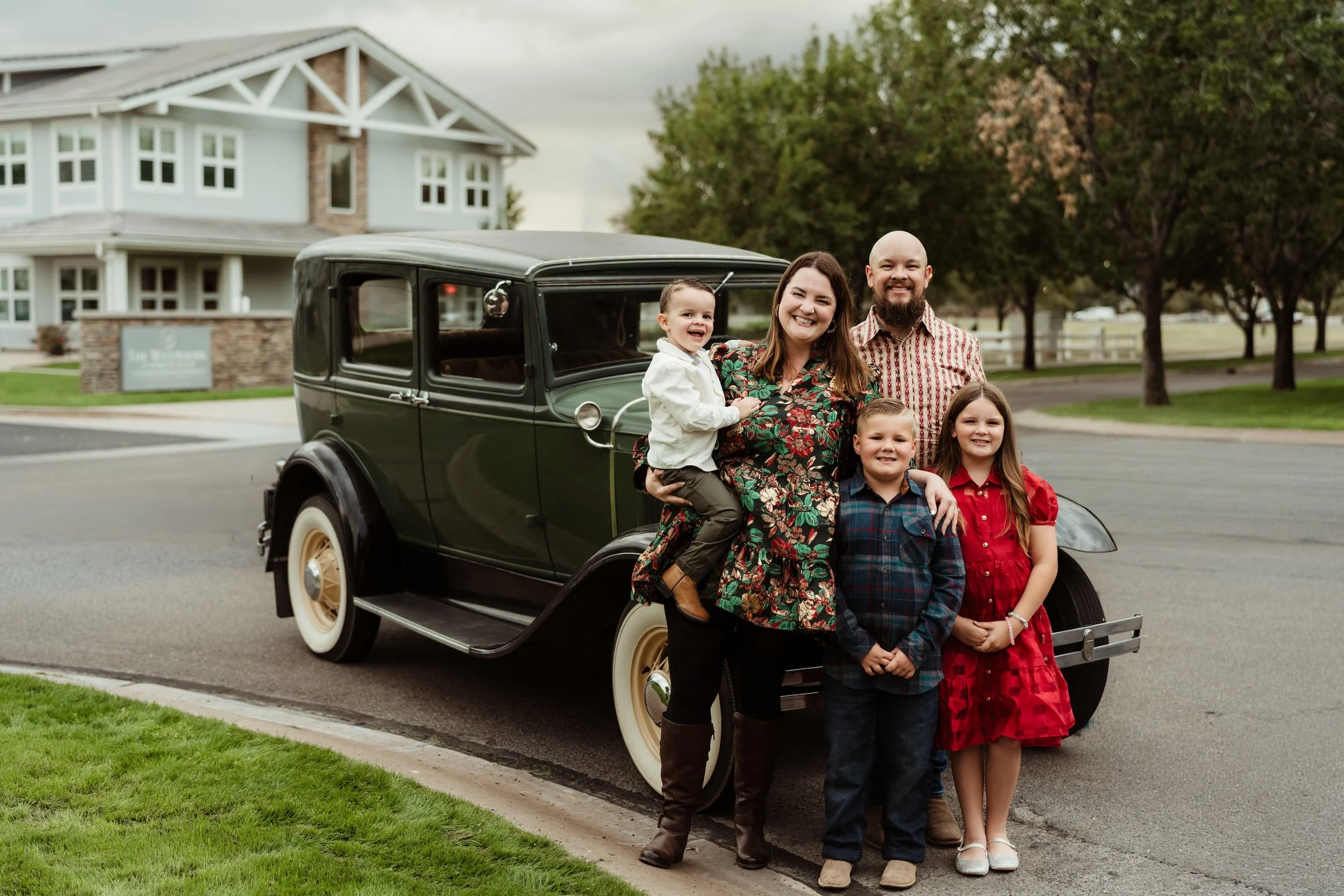 A Gabby Lane Classics family photo of five posing in front of a vintage 1931 Ford Four Door car on a residential street with trees and a house in the background.