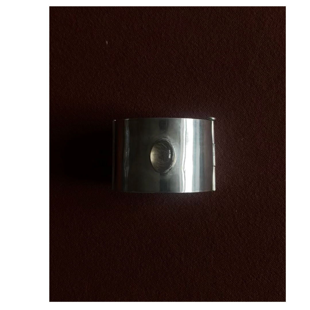 A wide silver cuff bracelet with a circular oval stone in the center, resting on a dark red textured surface.