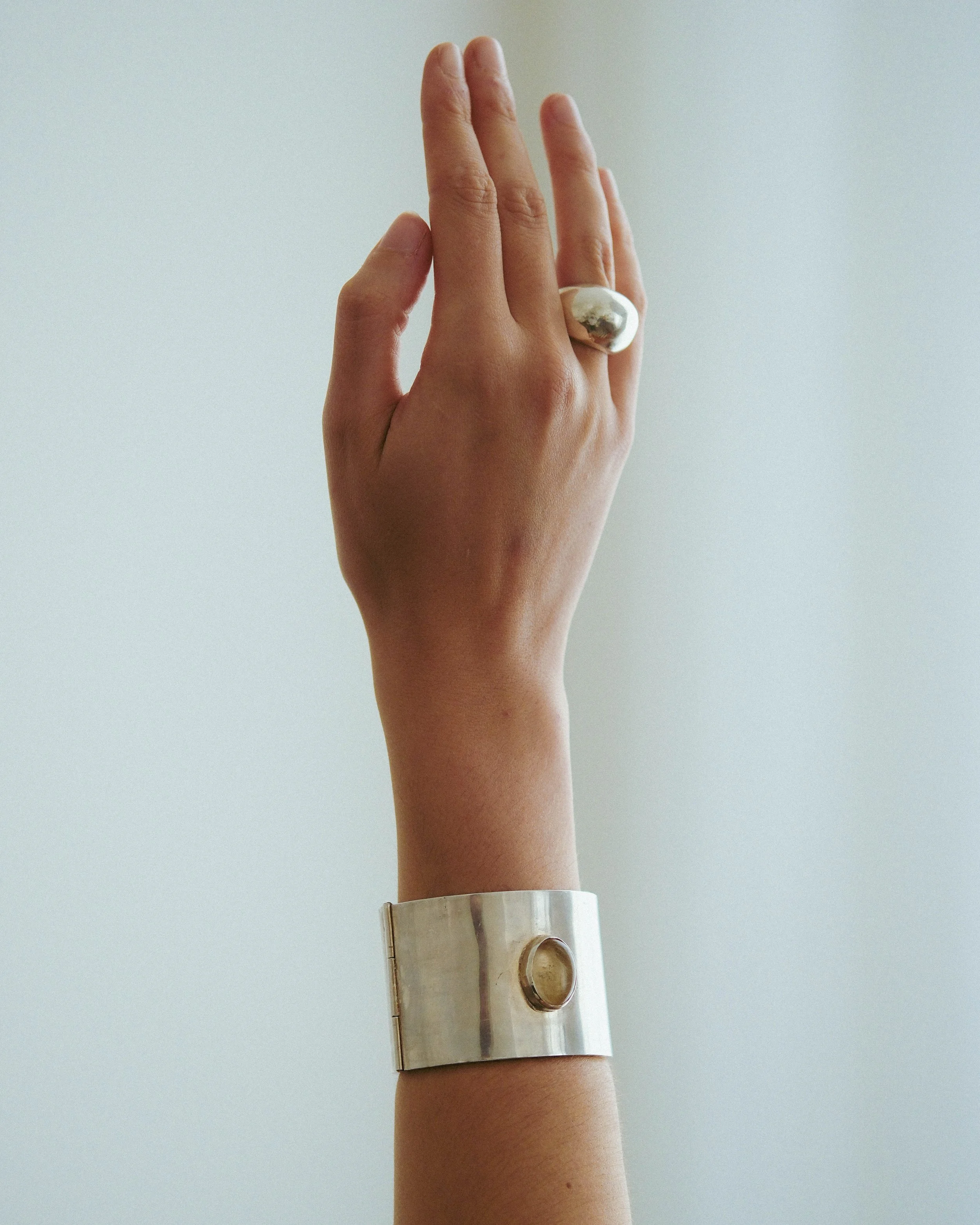 Close-up of a hand wearing a silver cuff bracelet with a large, irregularly shaped ring on the index finger, against a plain background.