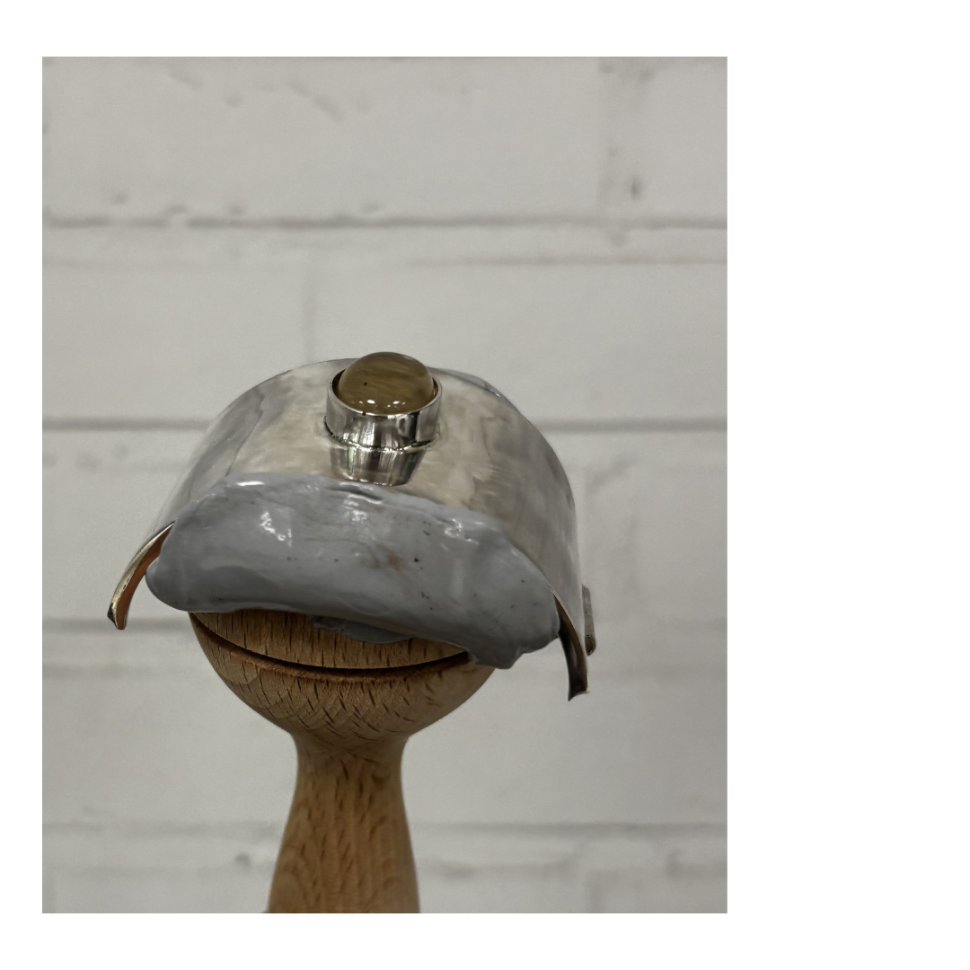 Object resembling a fish, made from a metal cap, a piece of white material, and mounted on a wooden stand, set against a light-colored wall background.