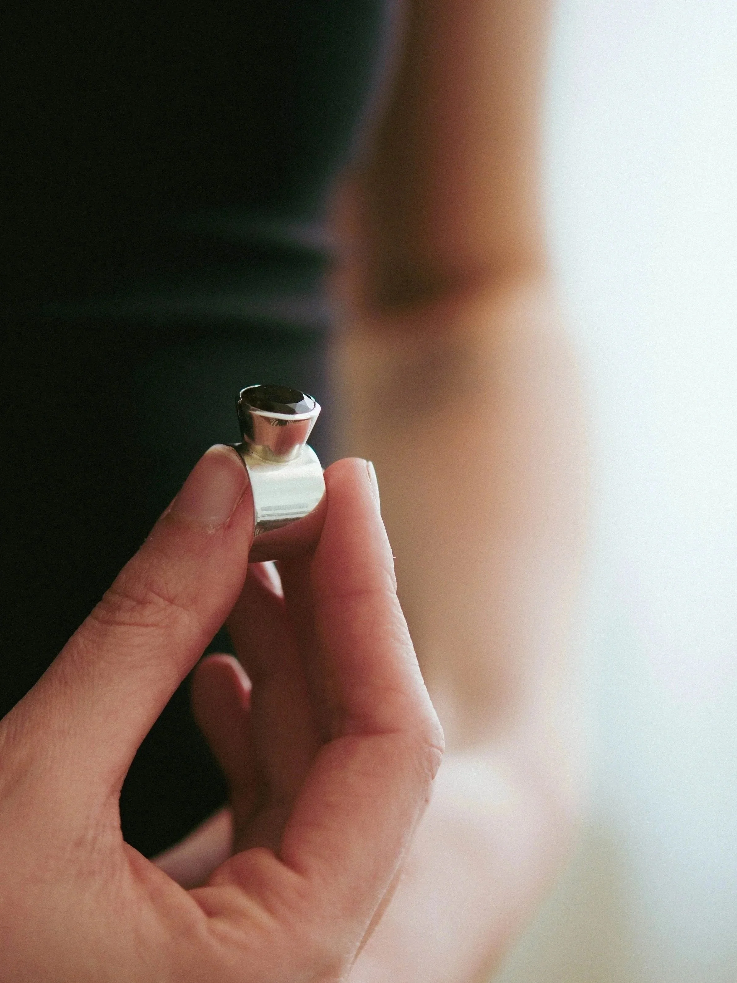 Close-up of a person's finger holding a silver ring with a black, shiny surface. The background is blurred with shades of dark and light colors.