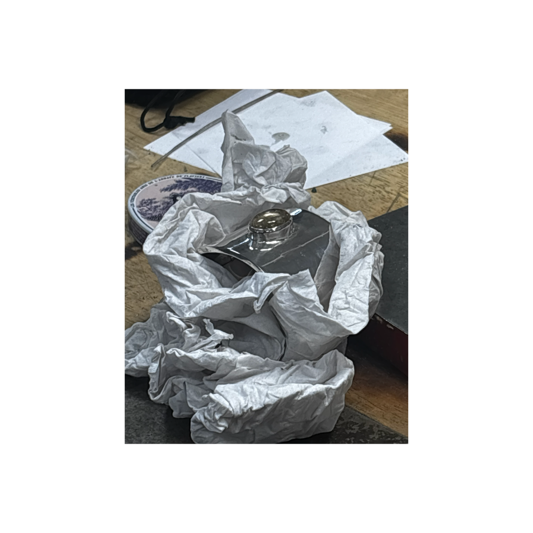 A crumpled piece of white tissue paper surrounds a metallic object, possibly a part of a watch, on a wooden table with papers, a tin, and other miscellaneous items scattered around.