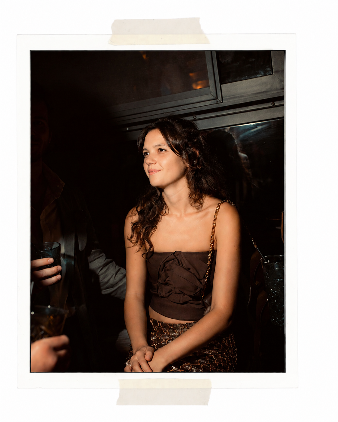 A young woman with curly brown hair wearing a brown crop top with spaghetti straps and patterned pants, standing and smiling in a dimly lit room, holding hands in front of her.