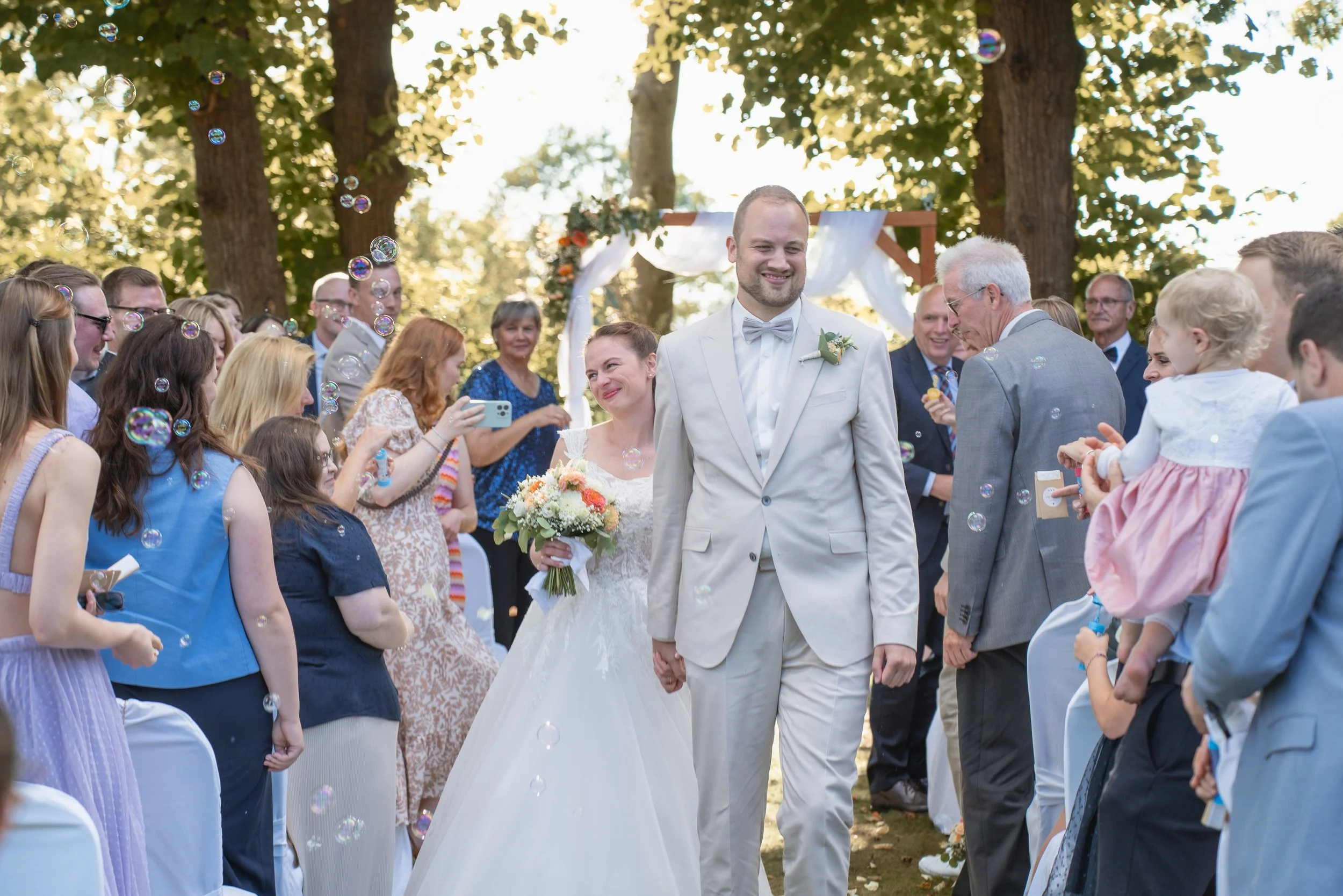 Hochzeit im Freien mit Braut und Bräutigam, umgeben von Gästen, im Sonnenschein zwischen Bäumen, mit Seifenblasen im Bild.