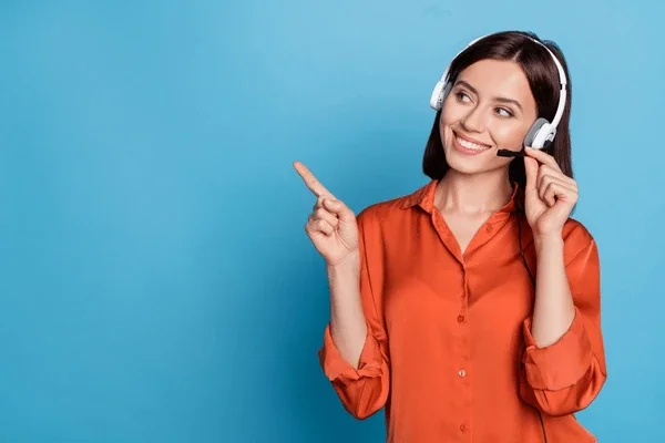 Smiling woman with a headset pointing to the side, wearing an orange blouse against a blue background.