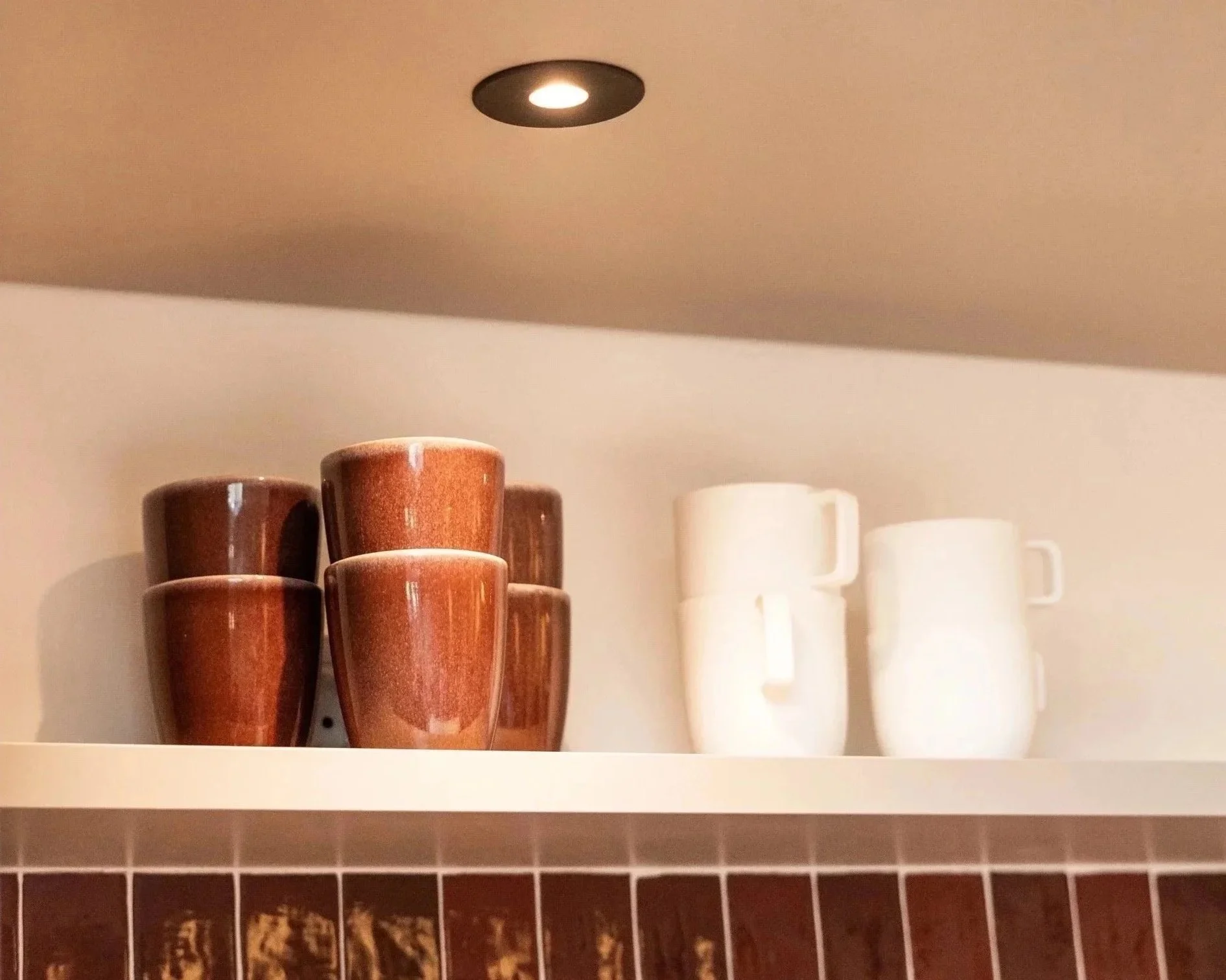 Twee groepen kopjes, één groep van bruin keramiek en één groep van wit keramiek, staan op een witte plank in een keuken met een keramieken muurtje.