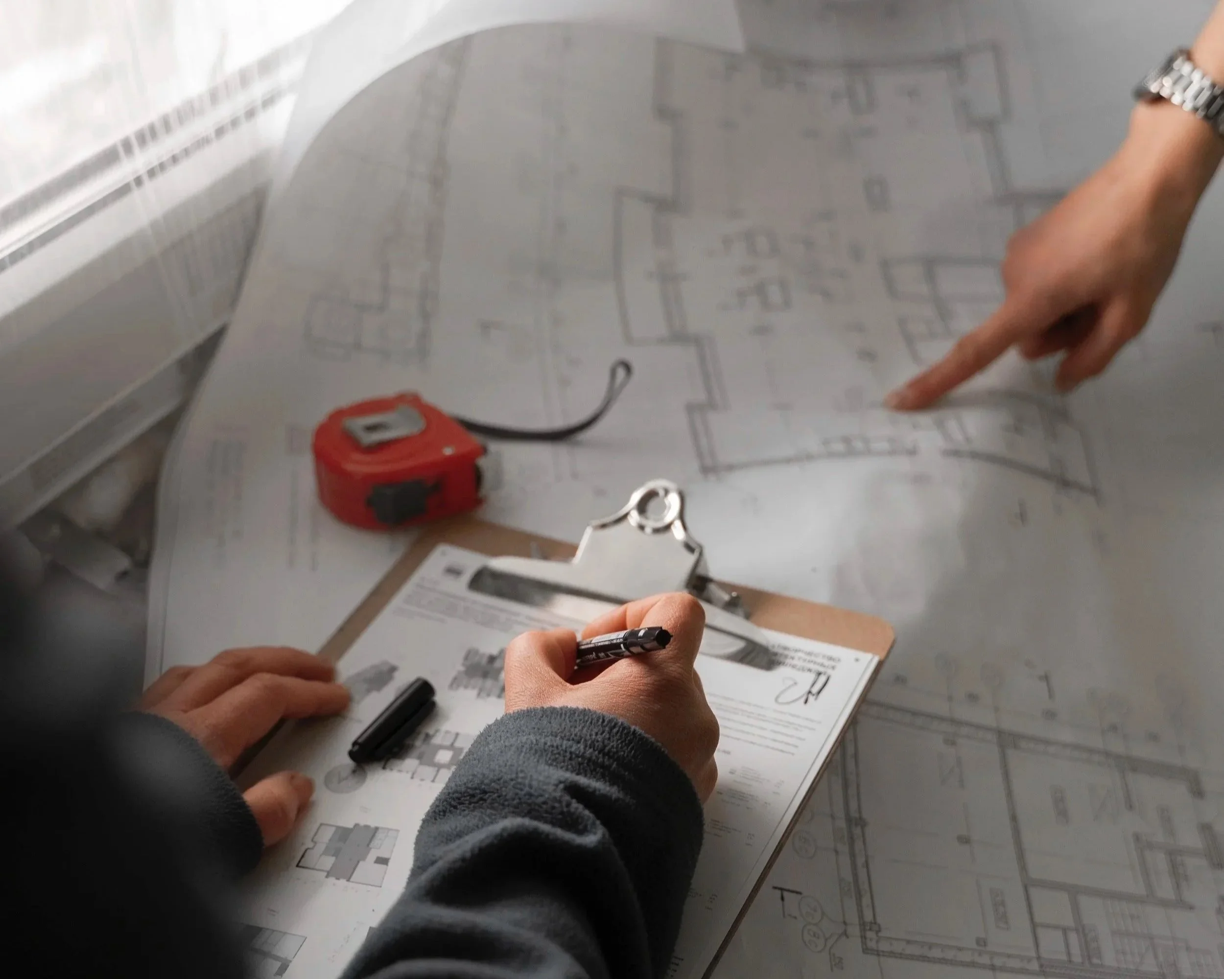 Twee mensen bekijken en markeren bouwtekeningen op een tafel, met meetlint en documenten.