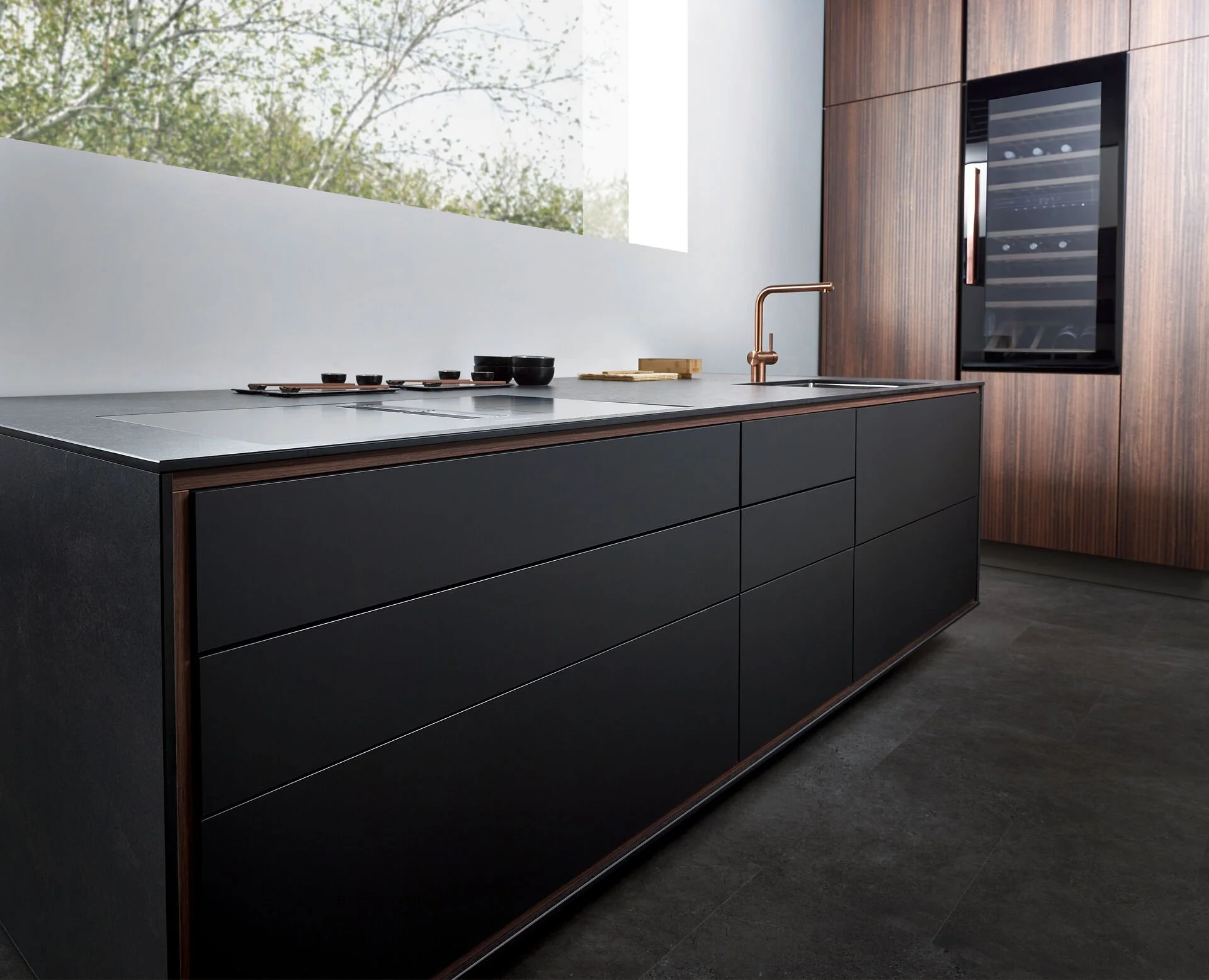 Moderne keuken met zwarte laden, houten wandkast en ingebouwde wijnkoelkast, wit aanrechtblad en grote ramen met uitzicht op bomen.