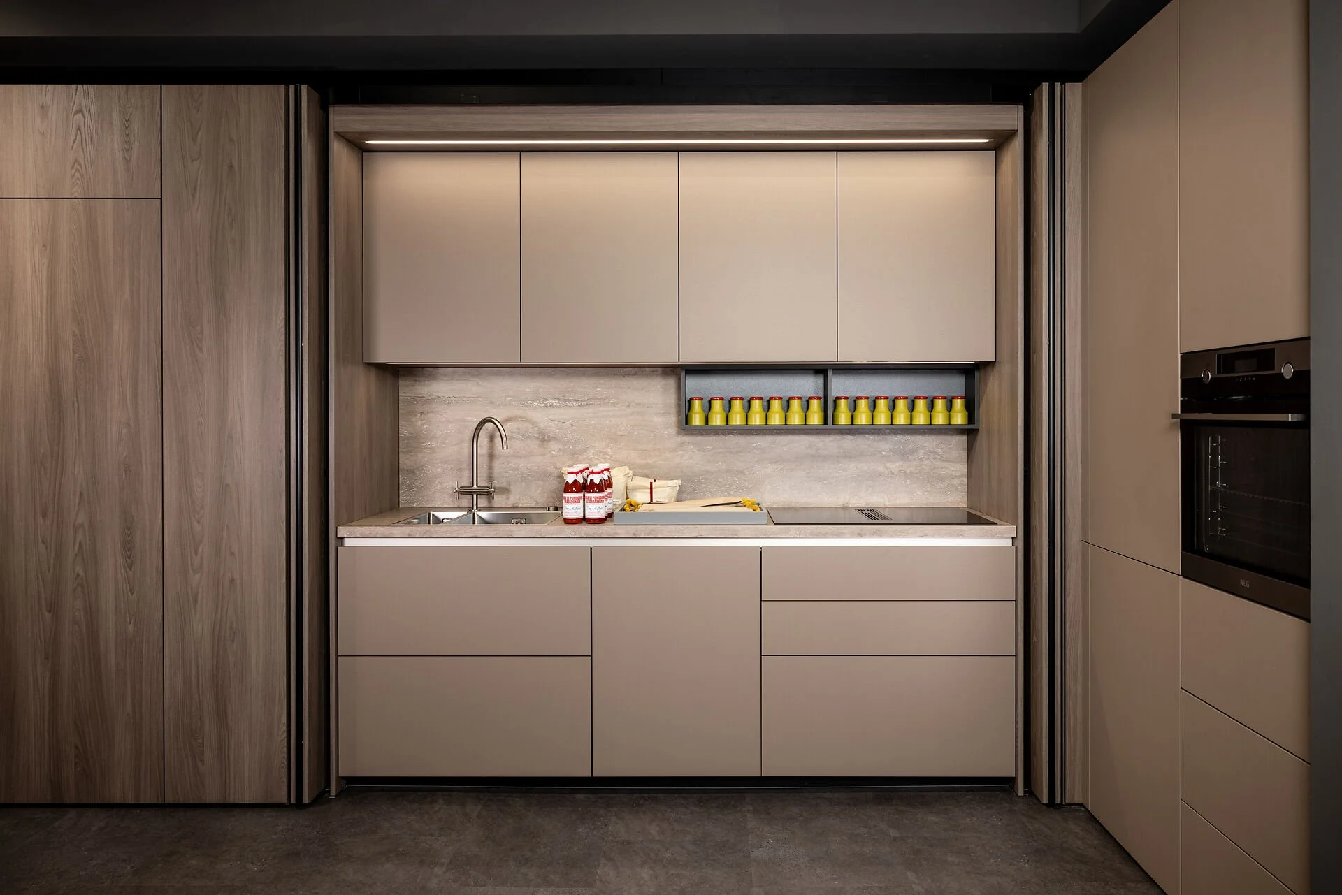 Moderne keuken met beige kasten, een wastafel, en een oven. Decoratieve gele flessen op een open plankenrek.