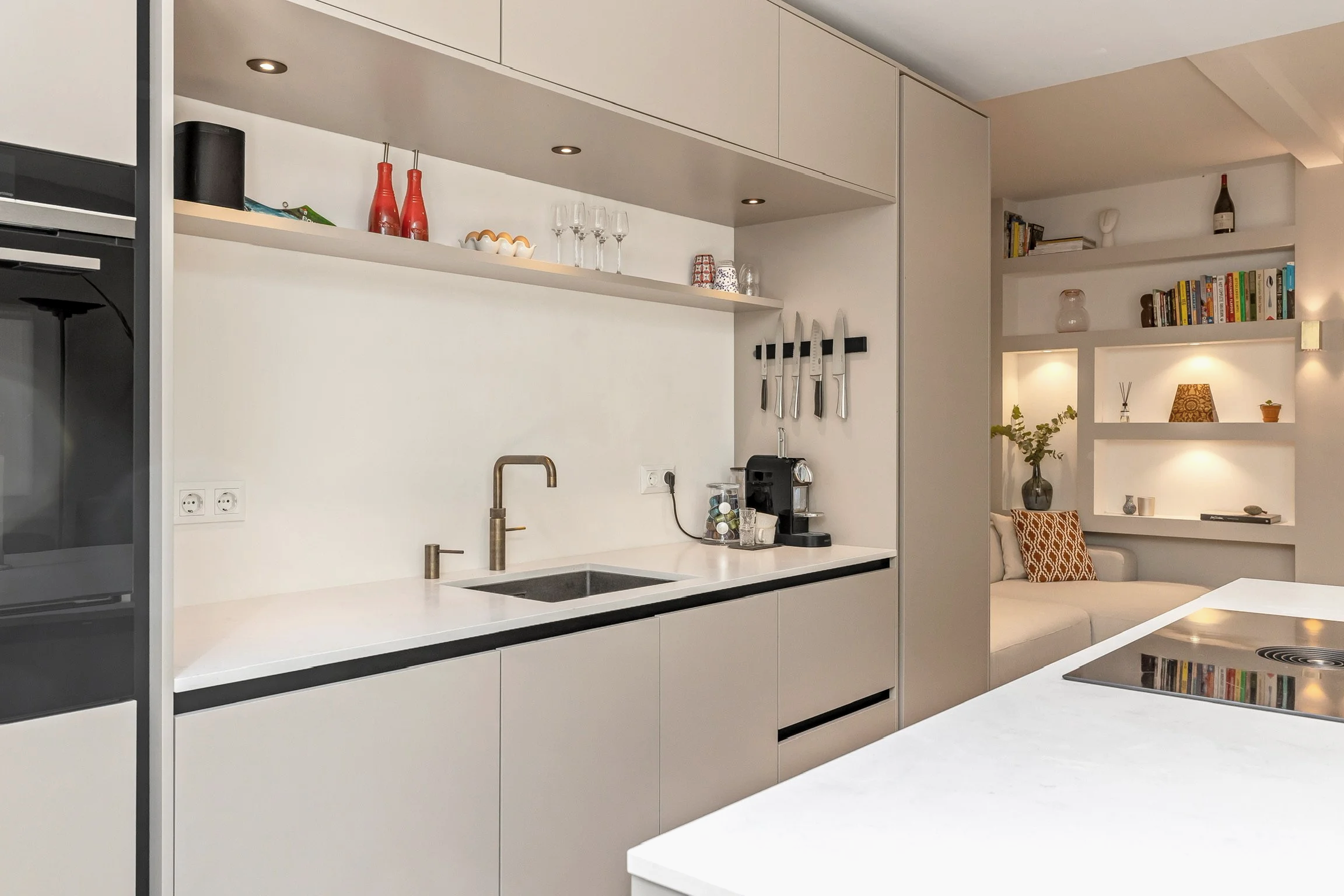 Moderne keuken met witte kasten, een koelkast, een gootsteen, een drip koffiezetapparaat, messen aan de muur en een open boekenkast met boeken en decoratie in de woonkamer.