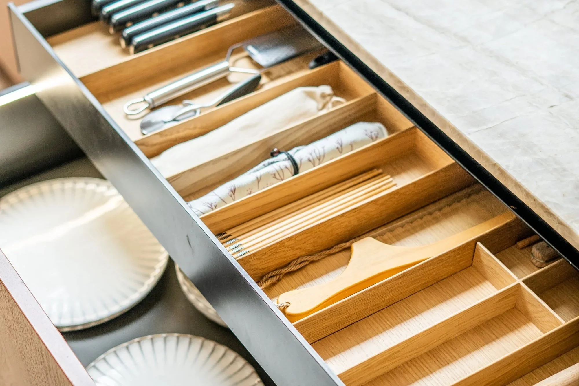 Open keukenkast met houten besteklade met verschillende keukenaccessoires en borden.