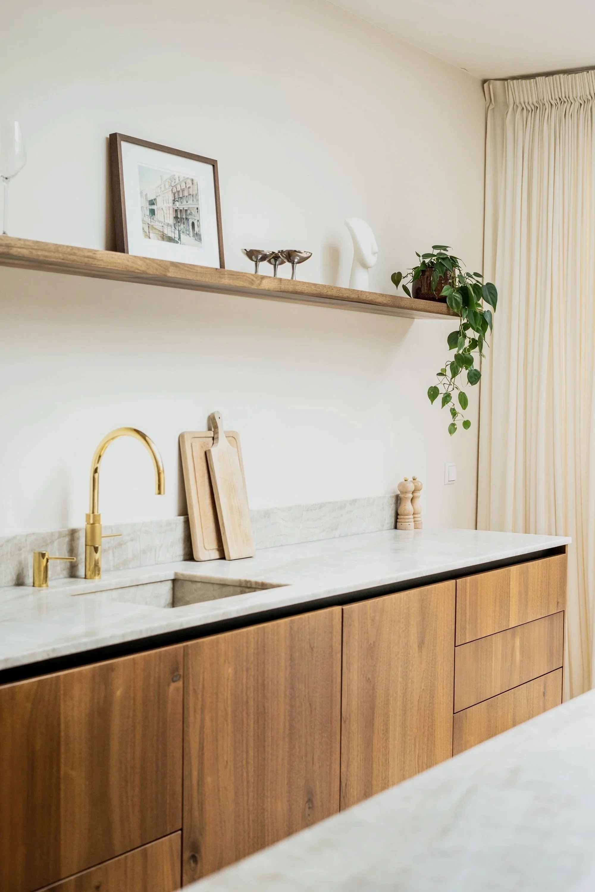 Moderne keuken met natuurlijke houttonen, gouden kraan, houten snijplanken, planten en decoratieve voorwerpen op een open houten plank, lichte gordijnen.