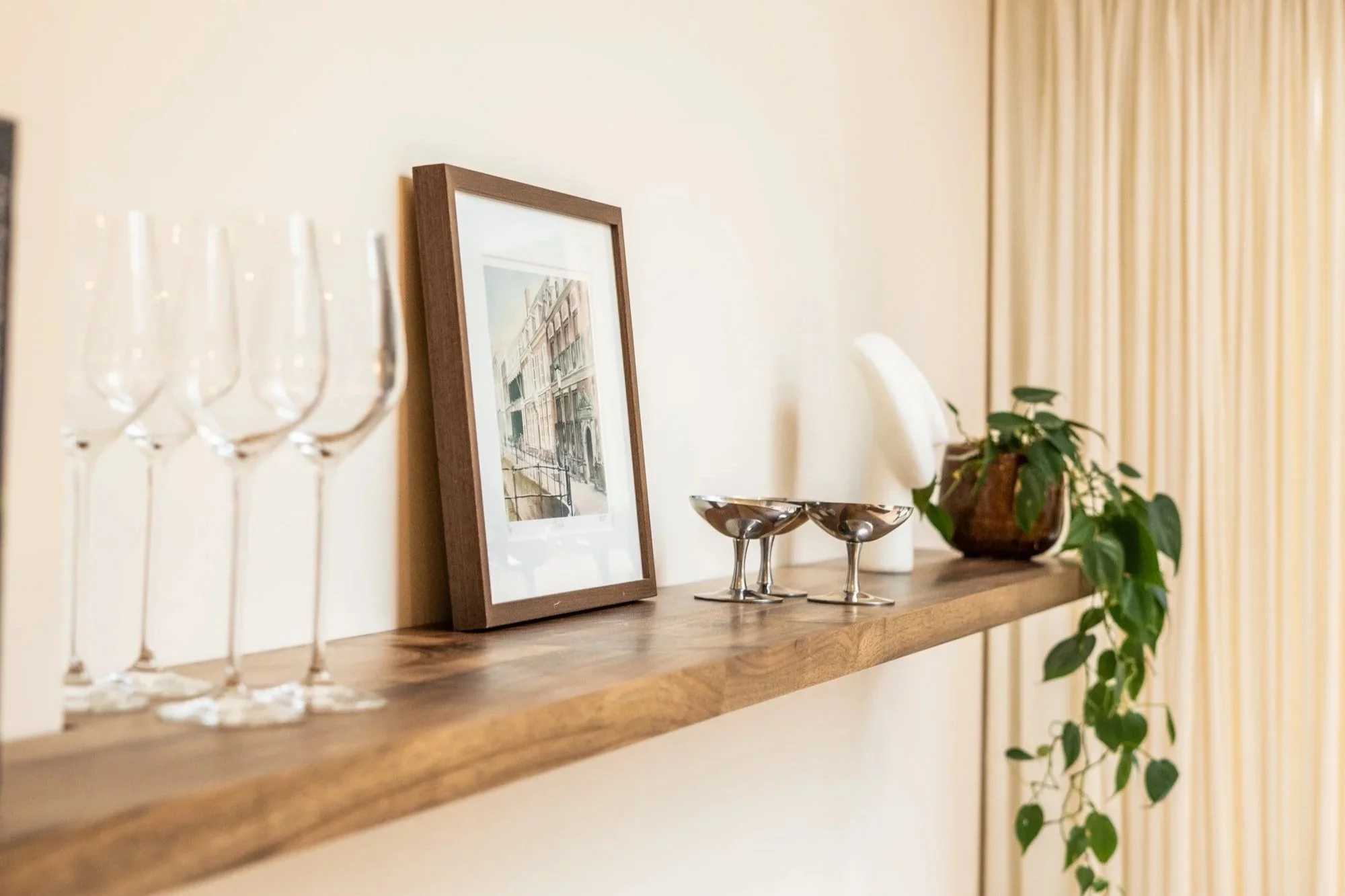Een houten boekplank met glazen, een ingelijste foto, twee metalen schaaltjes, een witte vaas en een plantenpot met groene bladeren, voor een beige muur met gordijnen op de achtergrond.