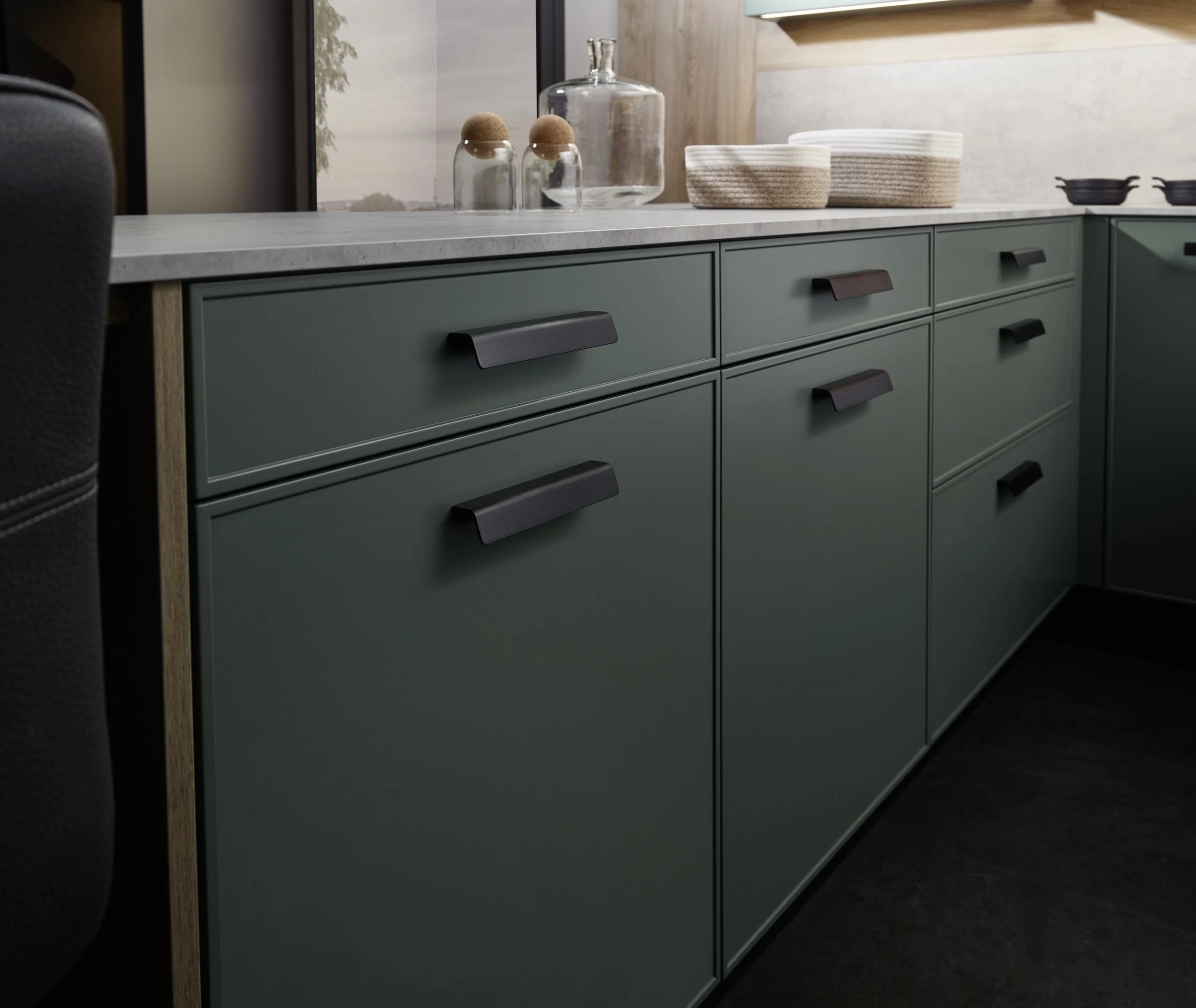 Groene keukenkasten met zwarte handvatten, witte stenen afvalbak op de keukentafel, glasfles, glazen potten en kommen op het aanrecht, uitzicht op deur met raam en buitenlandschap in de achtergrond.