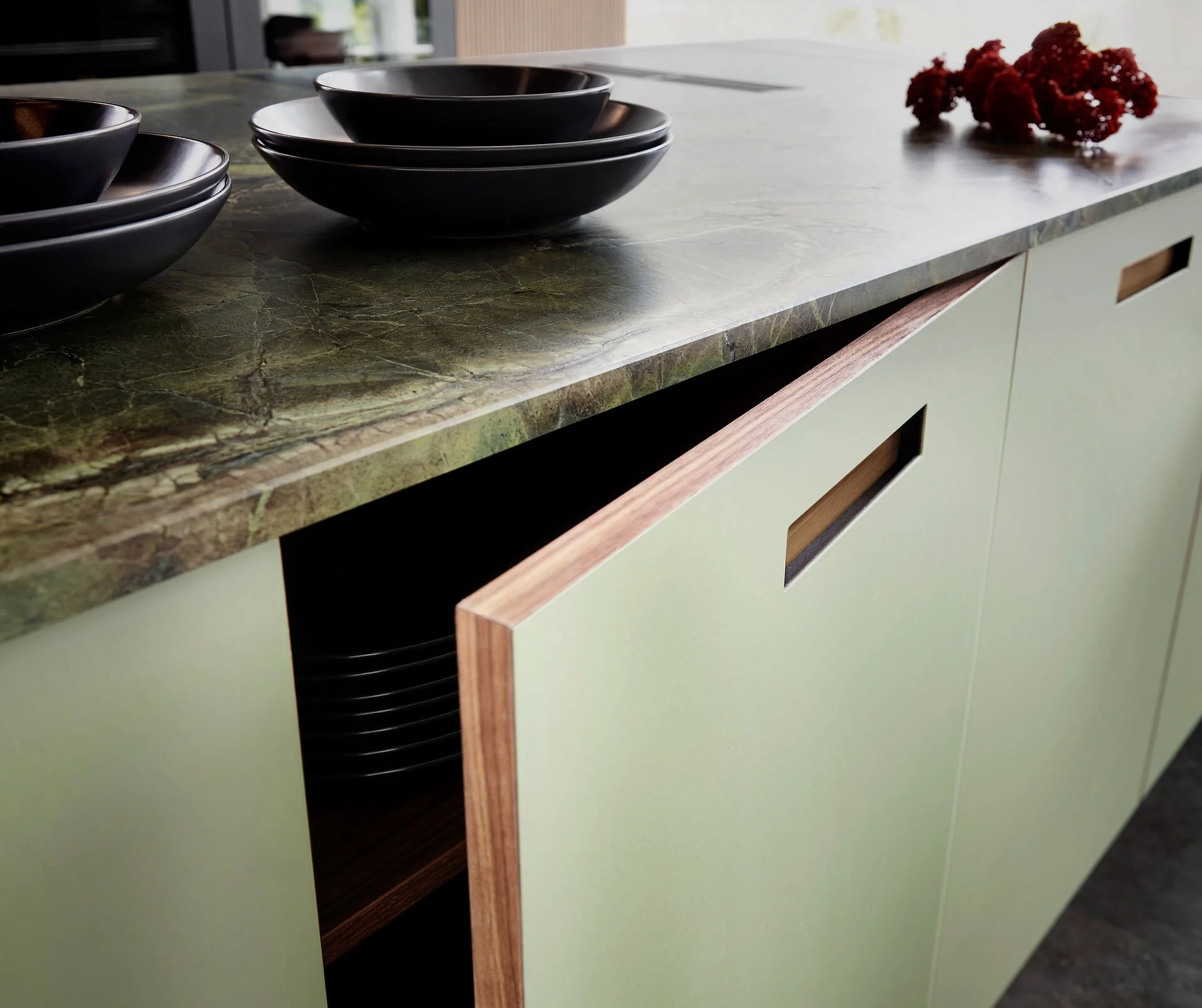 Keuken met groene kasten, een marmeren werkblad, en een deels open lade met zwarte borden, met enkele zwarte kommen op het werkblad en rode decoratie.