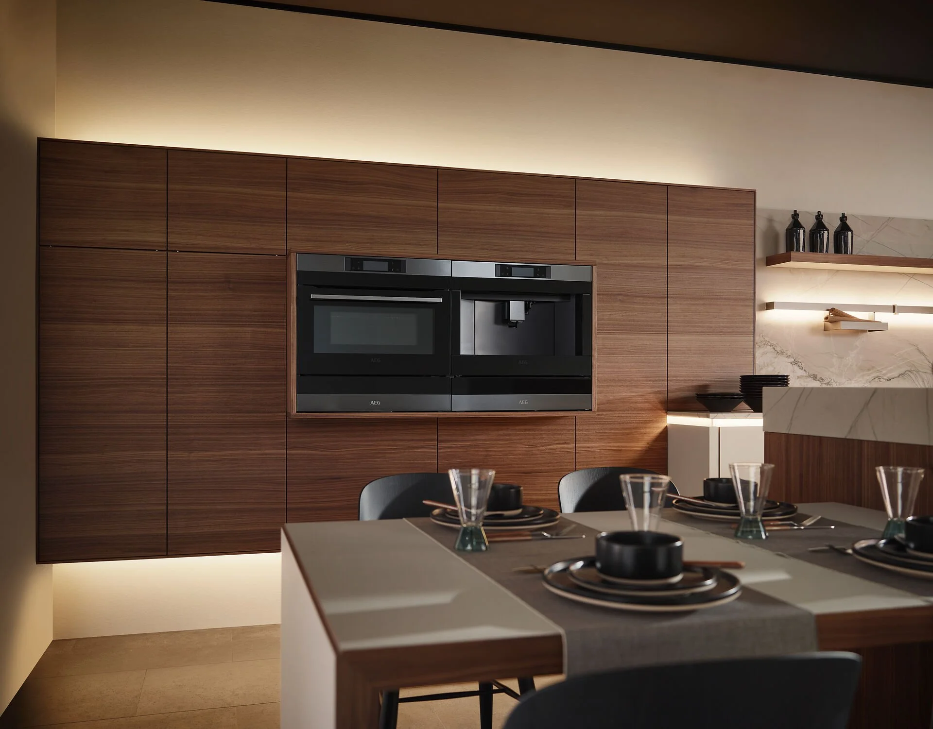 Een moderne keuken met een houten wandkast, ingebouwde oven en koffiezetapparaat, en een eettafel met tafelware en glazen.