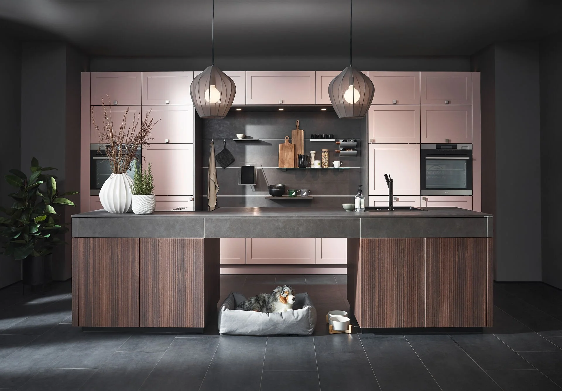 Moderne keuken met lichtroze bovenkasten, zwart werkblad, twee hanglampen, decoratieve planken, en een hond in een hondenbed onder het keukeneiland.