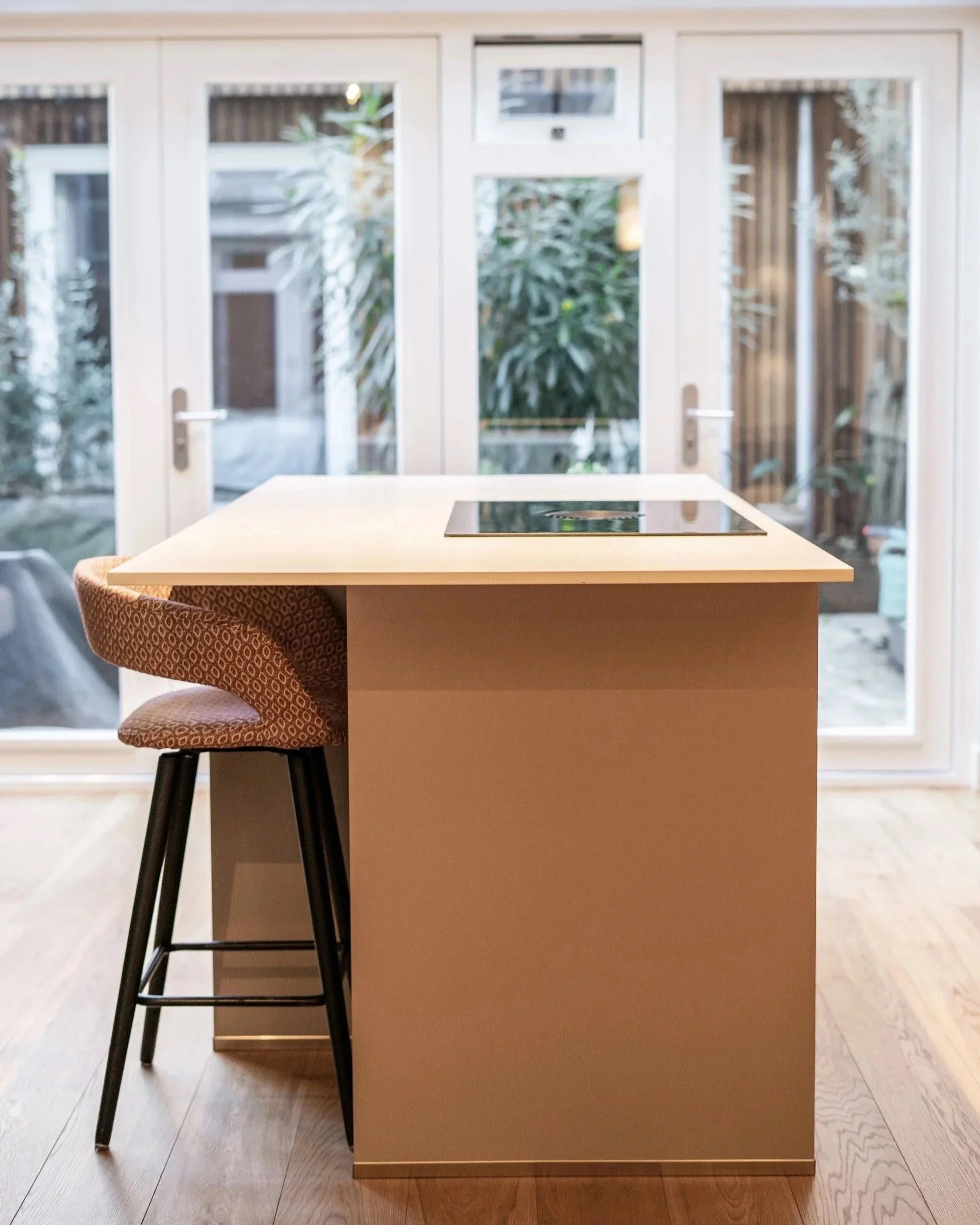 Een keuken met een eiland, een kookplaat en een stoel in een kamer met grote ramen en een tuin buiten.