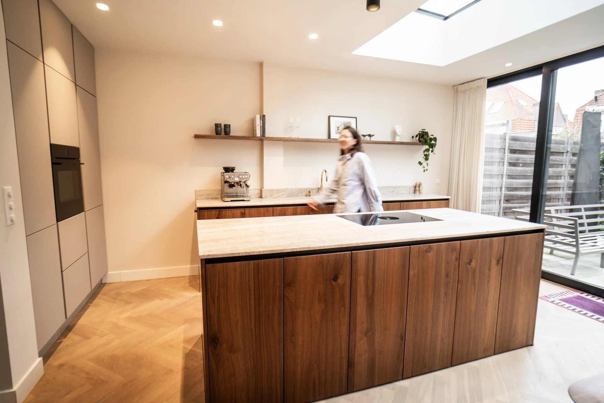 Moderne keuken met houten eilanden, een vrouw die erdoor loopt, en grote glazen schuifdeuren naar een tuin.