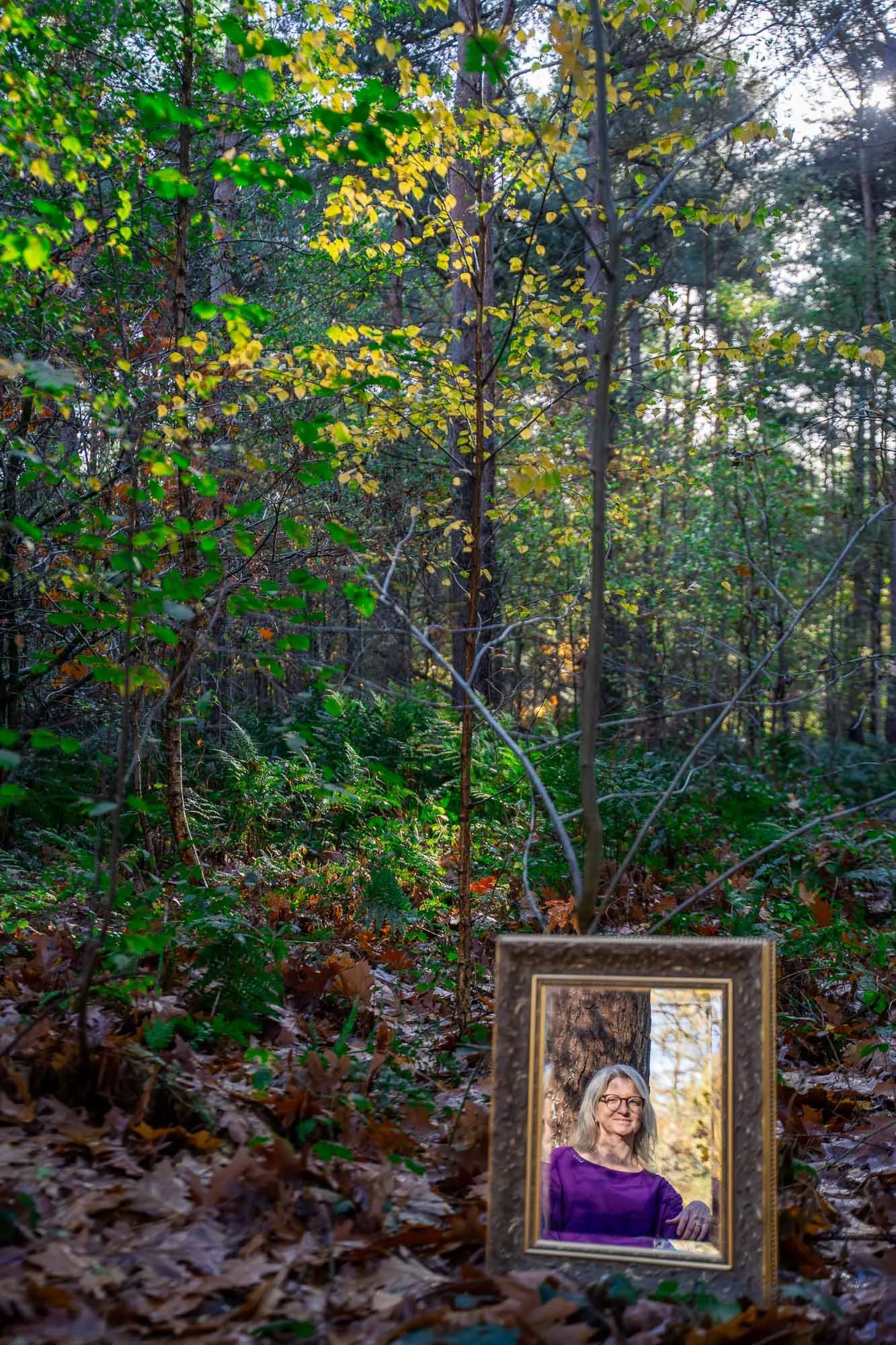 Een bos met groene en gele bladeren, en een spiegel tegen de boom waar je natuurcoach Marieke in ziet die in de spiegel kijkt.