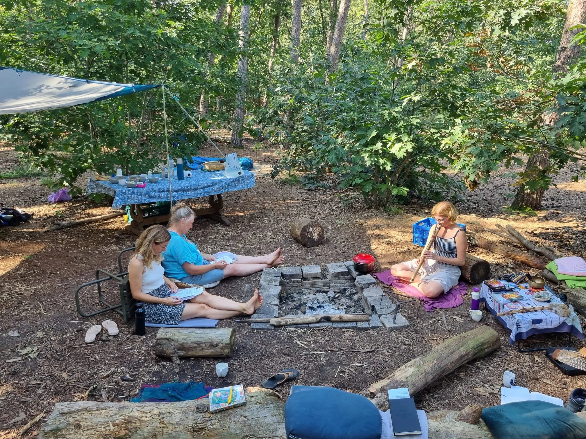 Drie vrouwen zitten op kussens rond een kampvuur in het bos, lezen en spelen hun instrumenten.