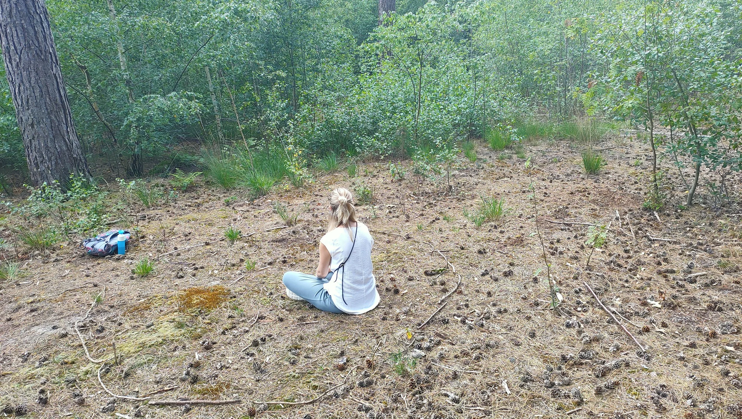 Persoon die op de grond zit in een bos, omringd door dennenappels en struiken, met een rugtas en waterfles naast zich.