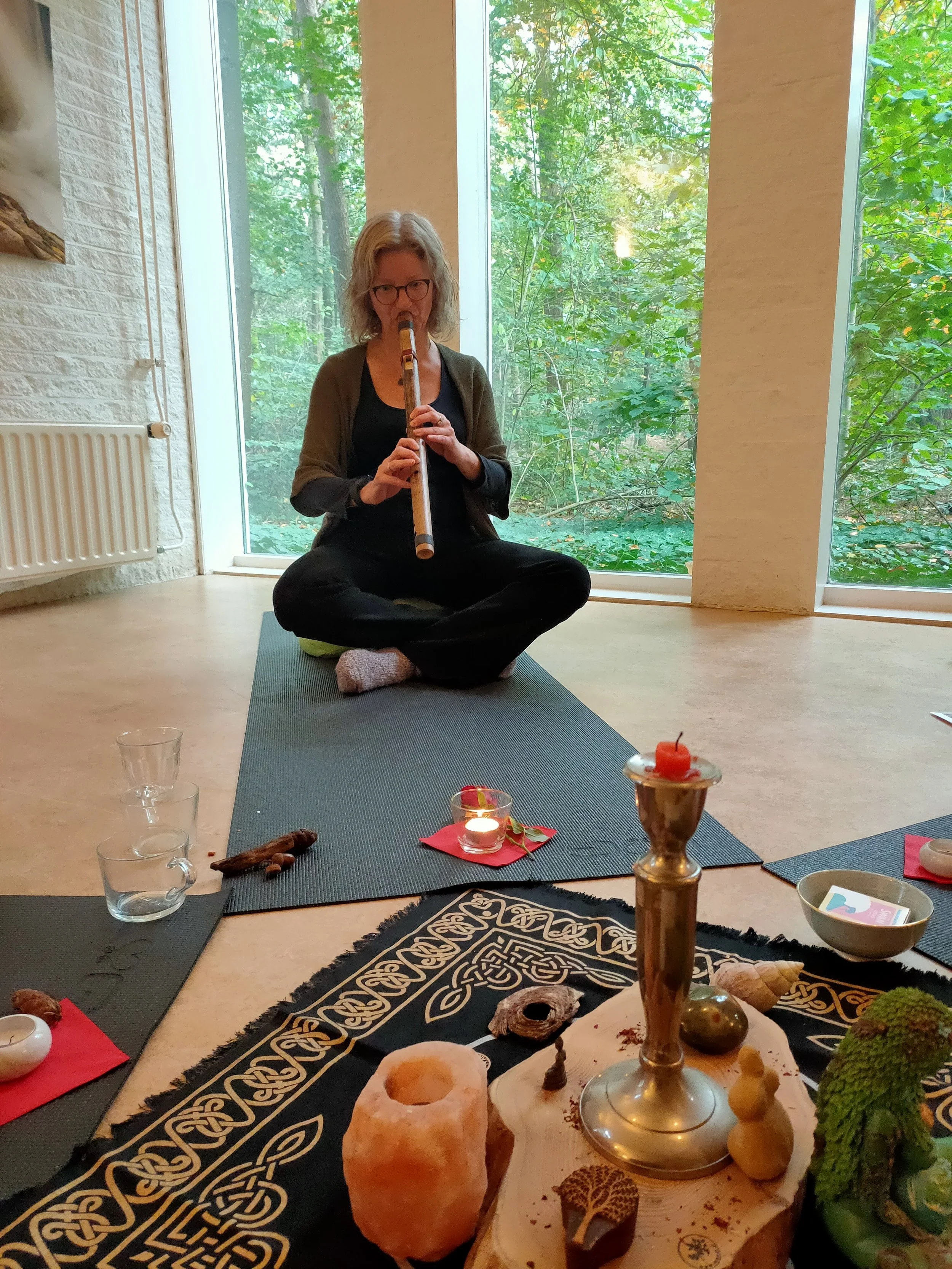 Natuurcoach Marieke zit gekruist op een matje in het Boshuis met grote ramen, terwijl ze op een indian love fluit blaast. Met een groen bos zichtbaar door de ramen.