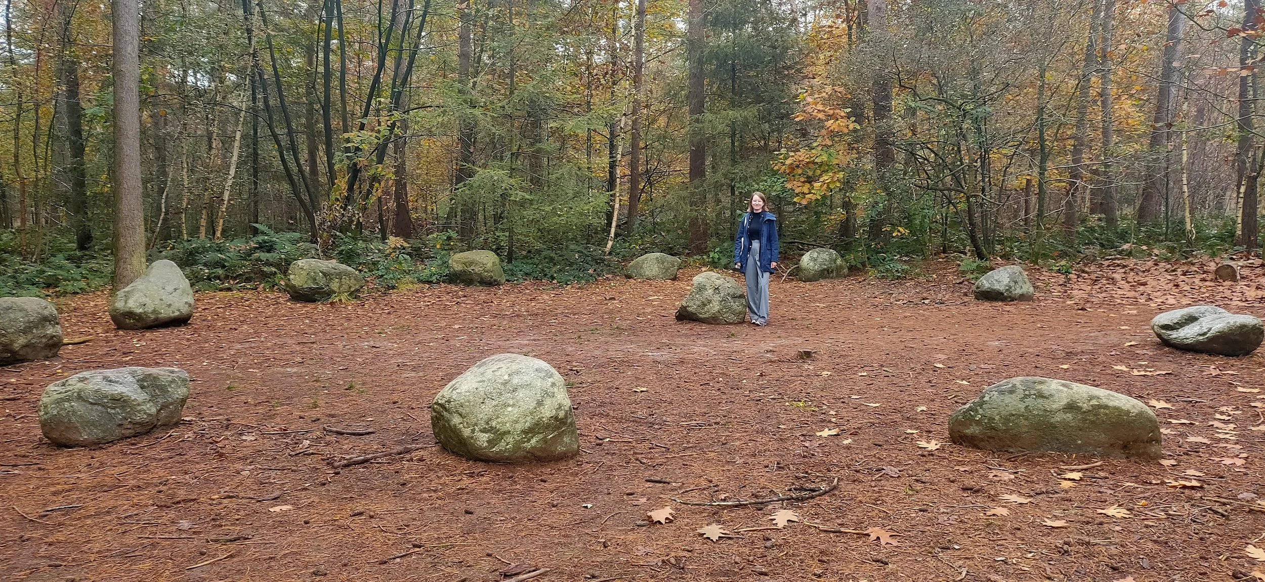 Vrouw staat in een bos, omringd door grote stenen, herfstkleuren in de bomen op de achtergrond.
