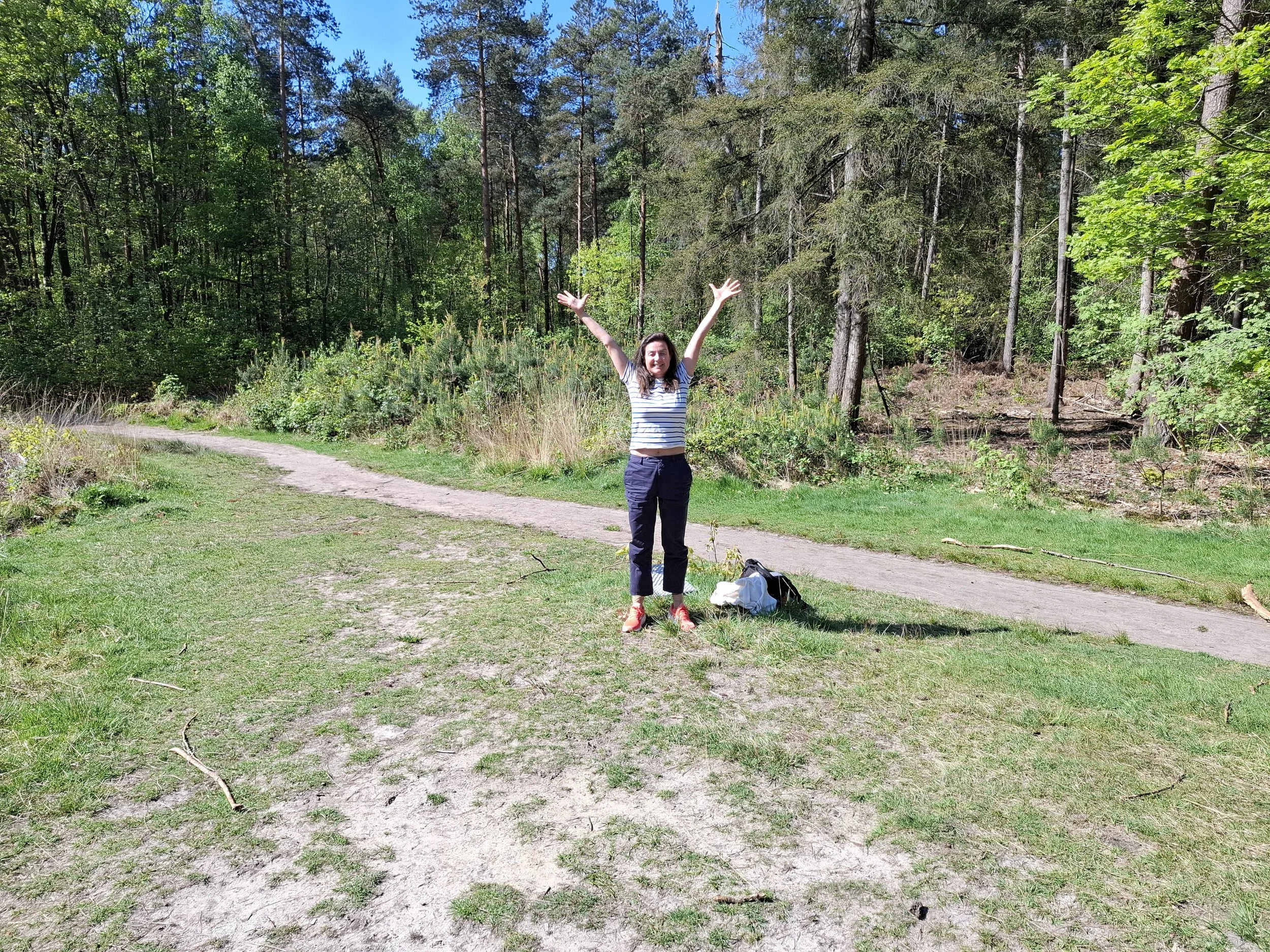 Vrouw met opgetrokken armen staat op een open plek in het bos, blije uitdrukking, zonnige dag.