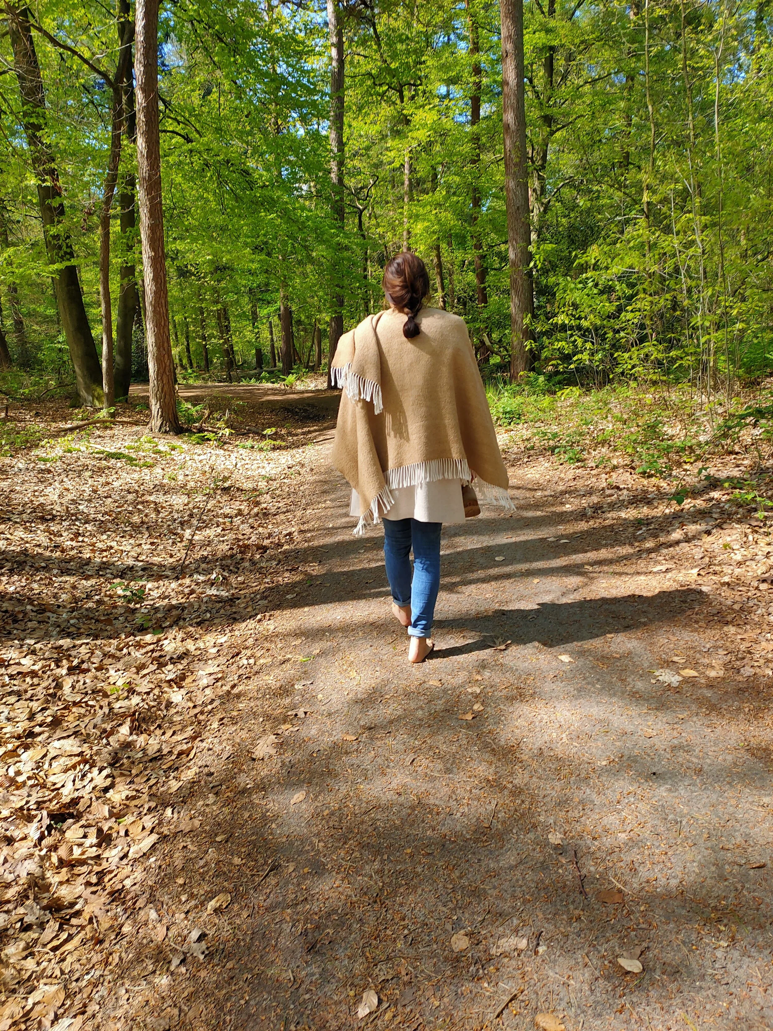 Een vrouw wandelt op blote voeten door het bos, gekleed in een beige jas en blauwe jeans.