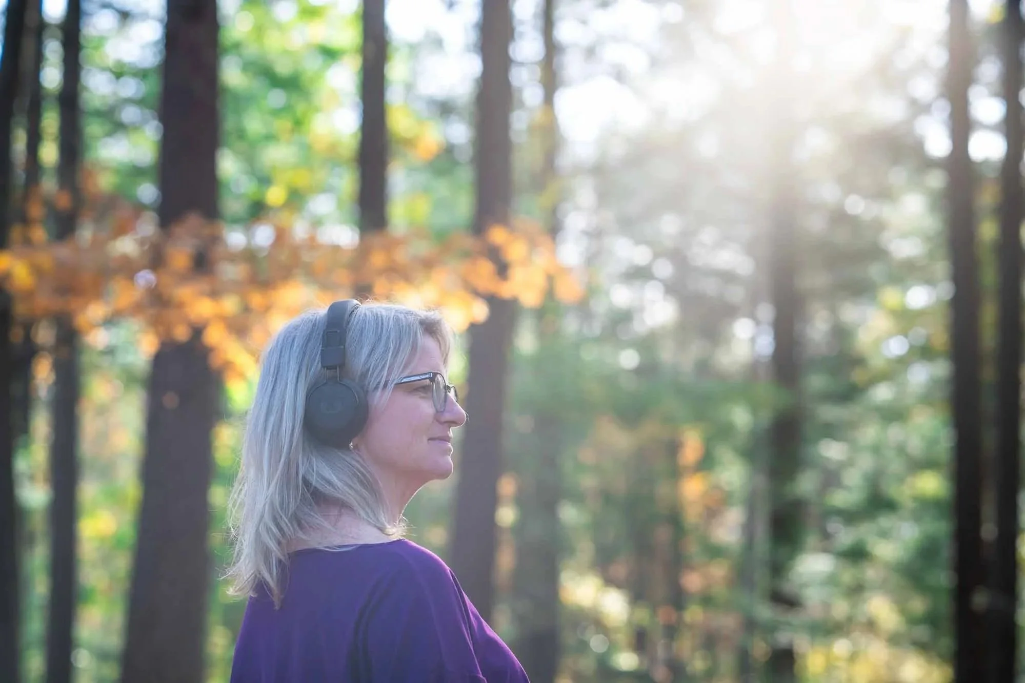 Marieke natuurcoach staat in het bos met koptelefoon en stralende zon tussen de bomen