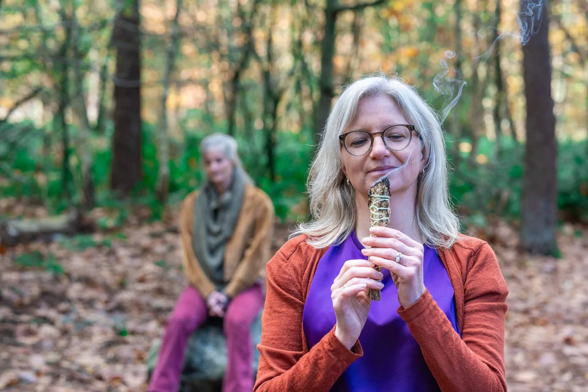Met een smudgestick doet natuurcoach Marieke een sjamanistisch ritueel in het bos met een coachee op de achtergrond zittend op een steen.