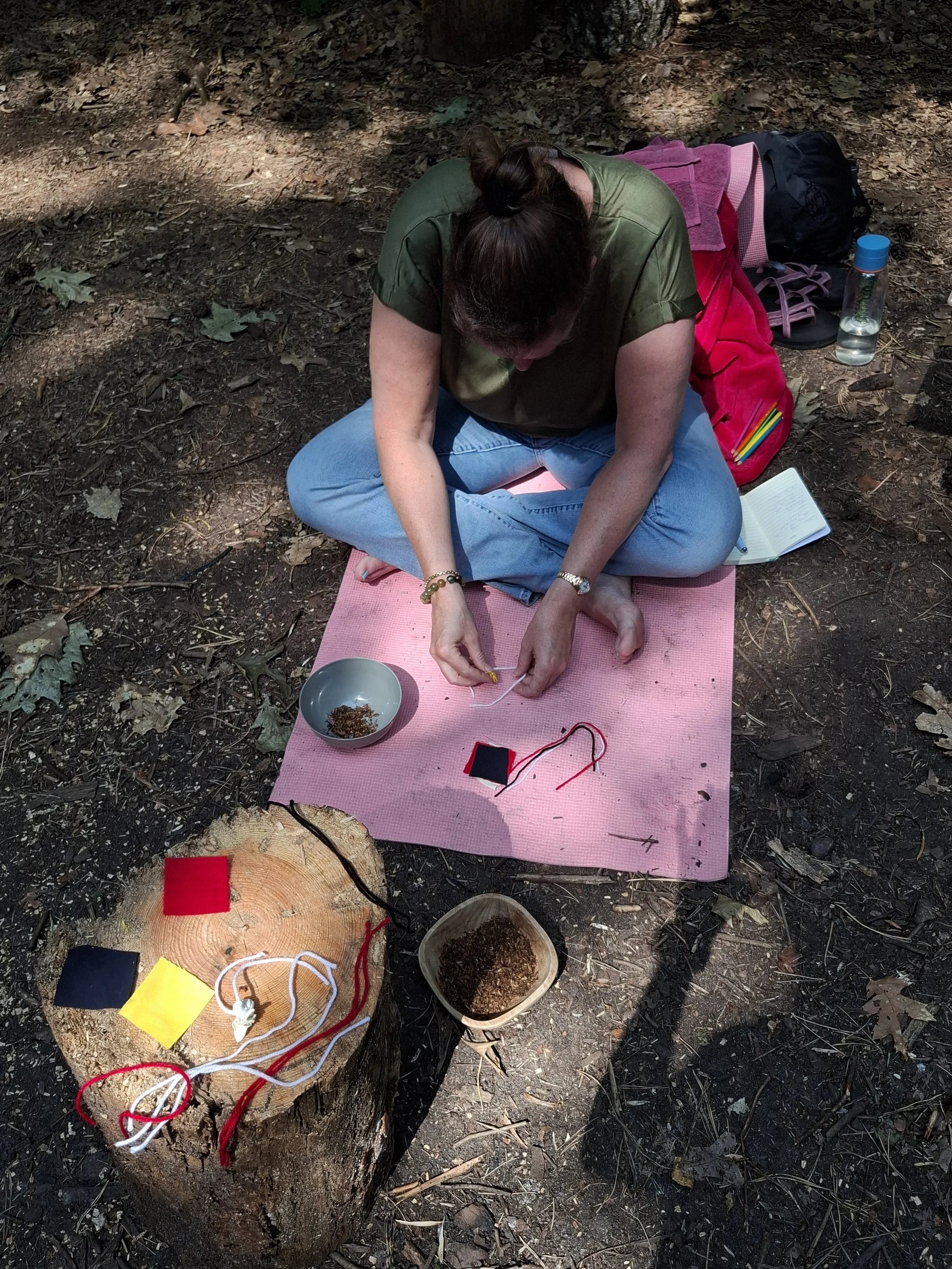 Vrouw zit op een roze matje in het bos en is bezig met het maken van sjamaistische instrumenten.