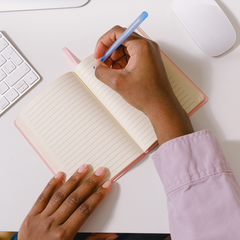 A person writing in a pink lined notebook with a blue pen, on a white desk with a white keyboard and mouse.