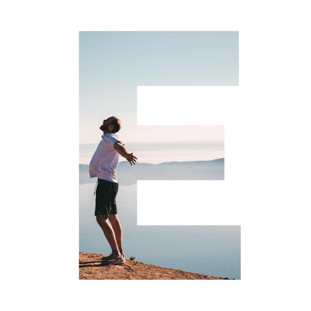A man standing on a sandy shore with arms outstretched, facing a calm lake during sunrise or sunset with hills in the background.