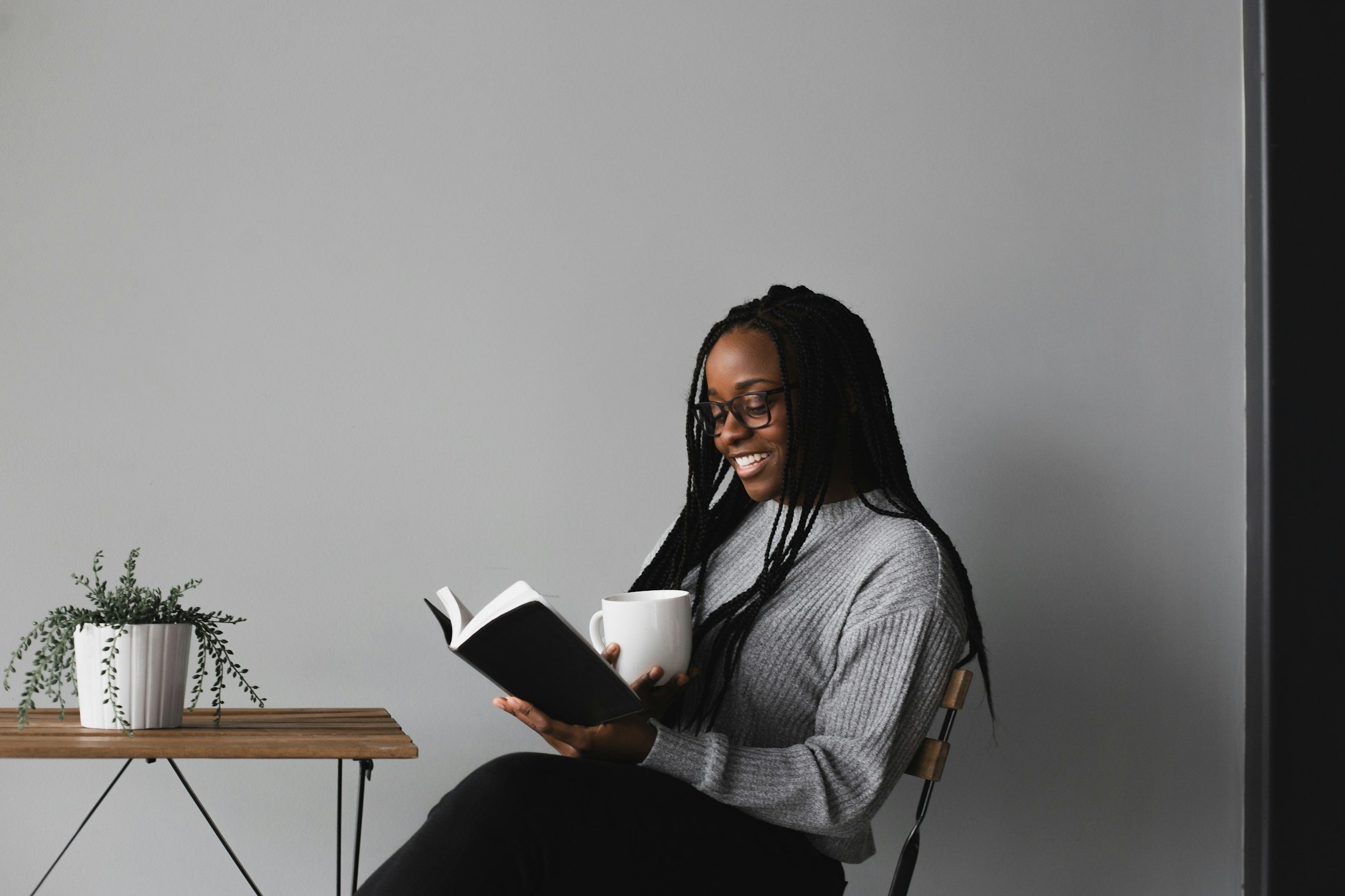 A woman with glasses and long braids sitting on a wooden chair, smiling and reading a book while holding a white mug, with a small potted plant on a wooden side table beside her against a plain gray wall.