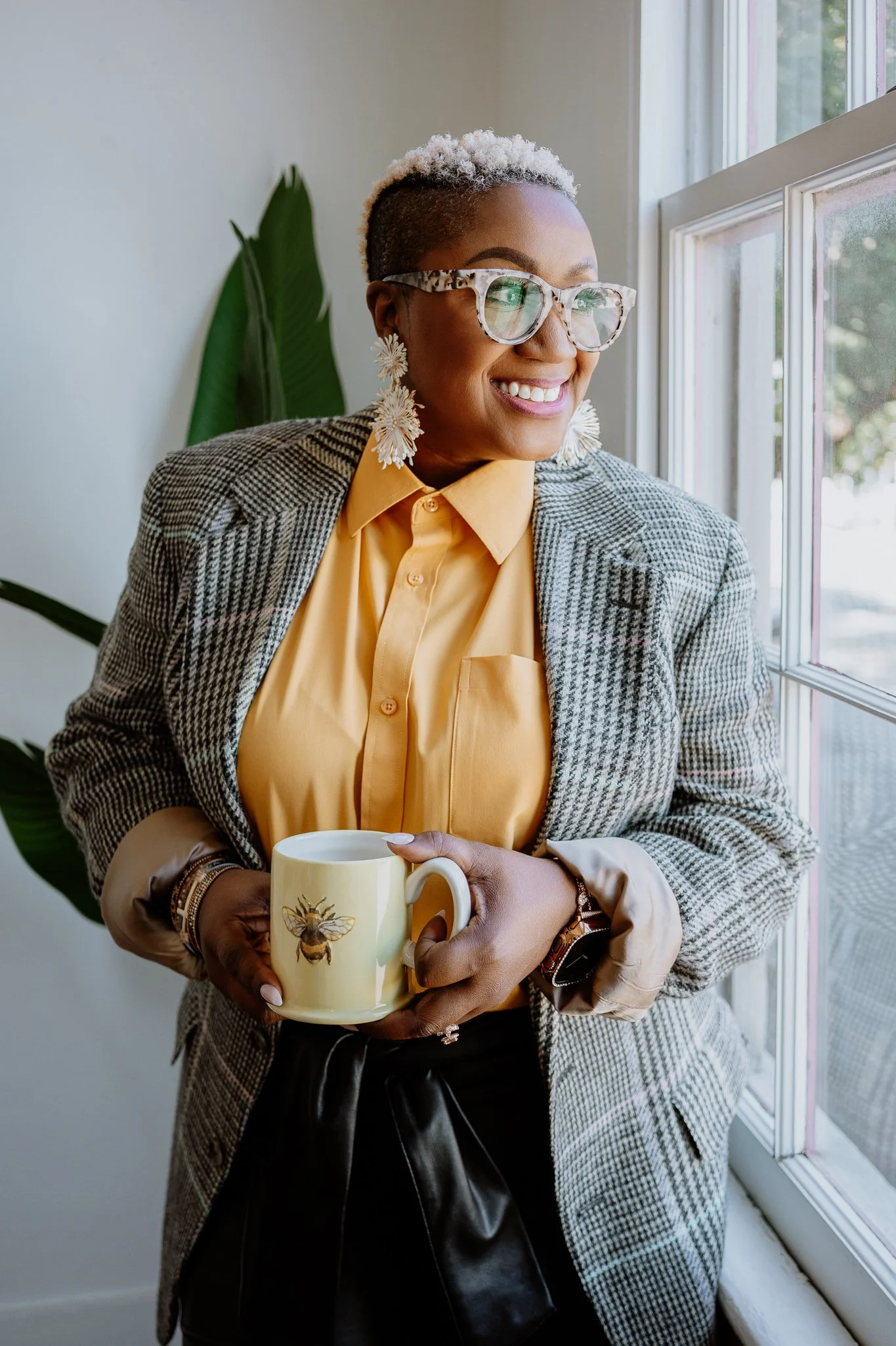 A woman with short blonde hair and glasses looking out a window, holding a beige mug with a honeybee illustration, wearing a yellow shirt, patterned blazer, and large floral earrings.