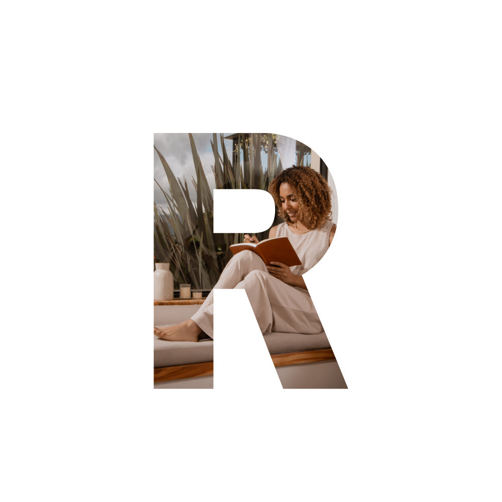 A woman with curly hair sitting on a window seat, reading a book, with a cozy indoor setting and plants in the background.