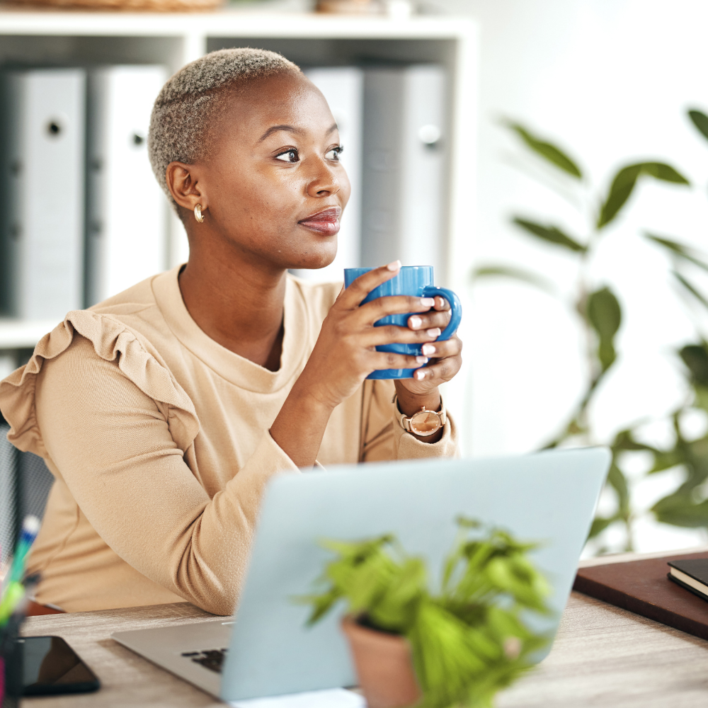 A woman with short blonde hair, wearing a beige top with ruffled shoulders, sitting at a desk holding a blue mug, with a laptop and green plant on the desk, in a bright office environment.