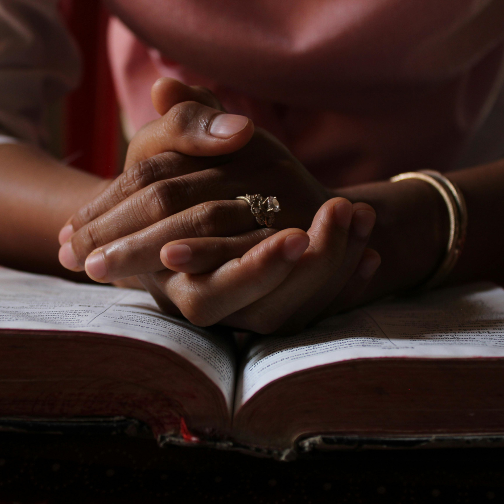 Close-up of intertwined hands resting on an open book, with one hand wearing rings and a bracelet, in a dimly lit setting.