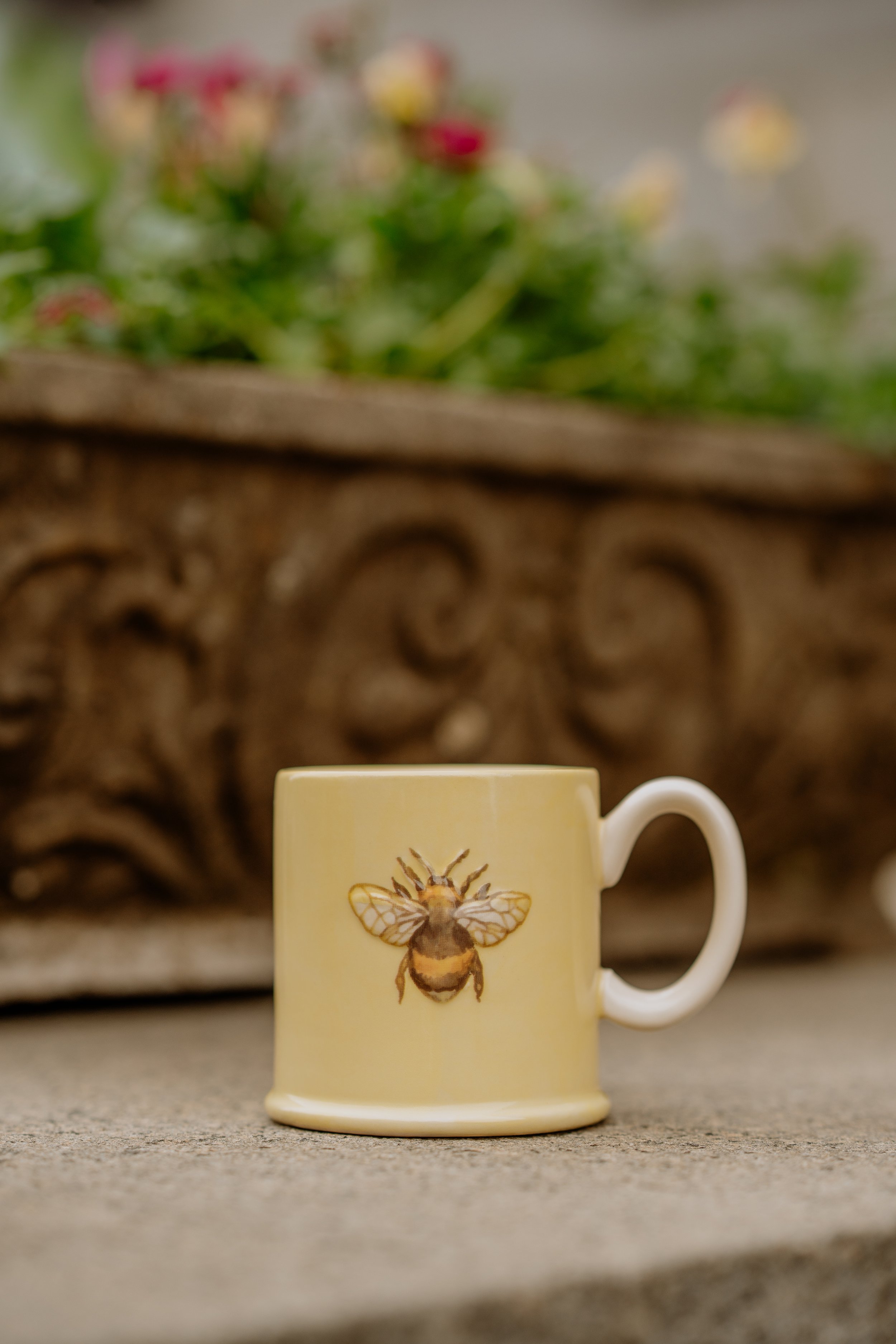 A beige mug with a bee illustration on the front, placed on a concrete surface with a blurred background of flowers and a decorative stone masonry.