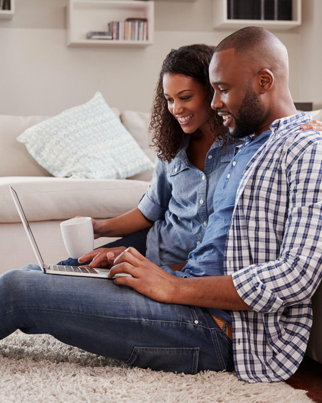A smiling couple sitting on the floor in front of a laptop, holding mugs, in a cozy living room.