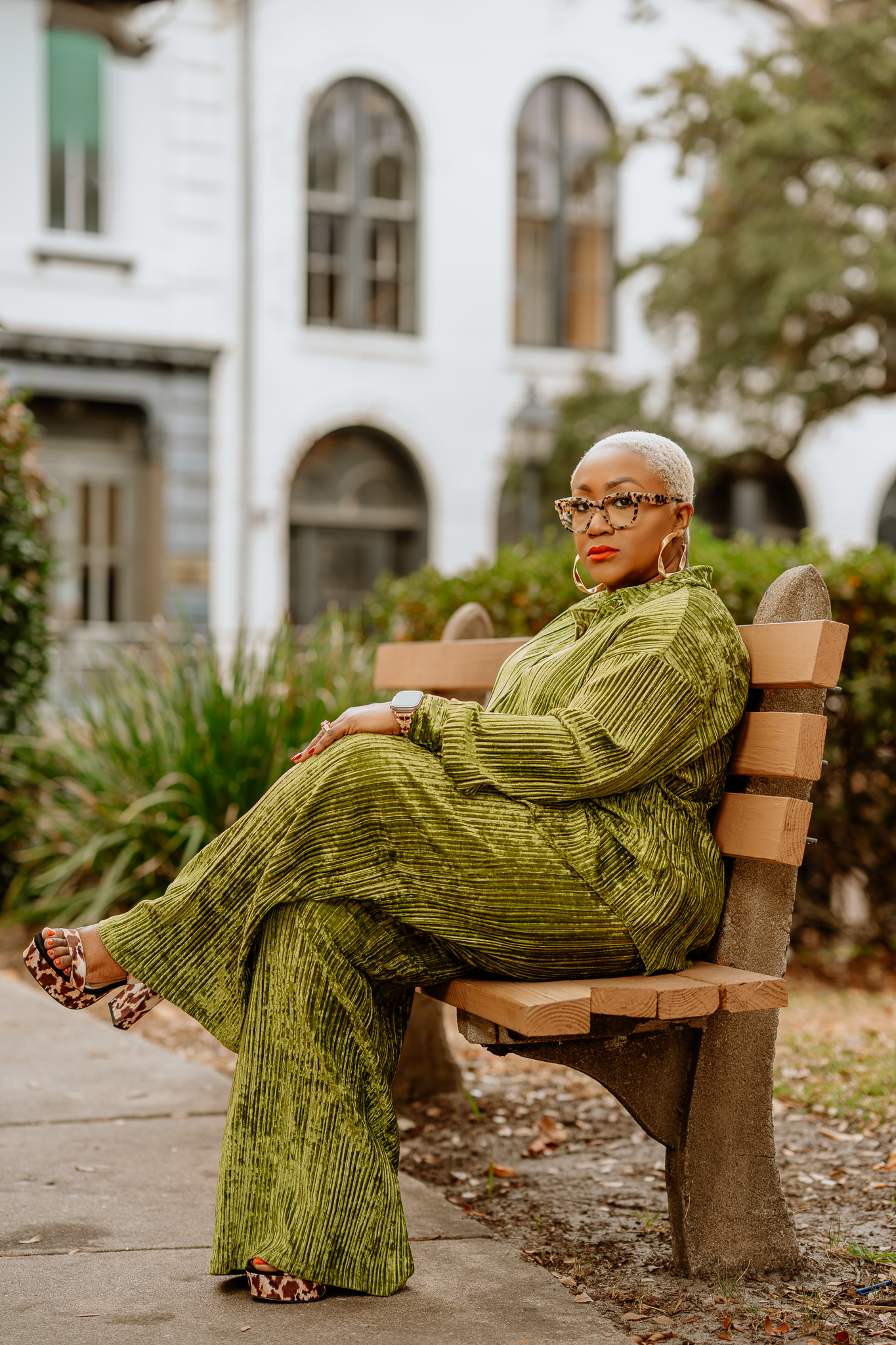 A woman with short platinum blonde hair, wearing glasses, earrings, and a green striped outfit, is sitting on a park bench.