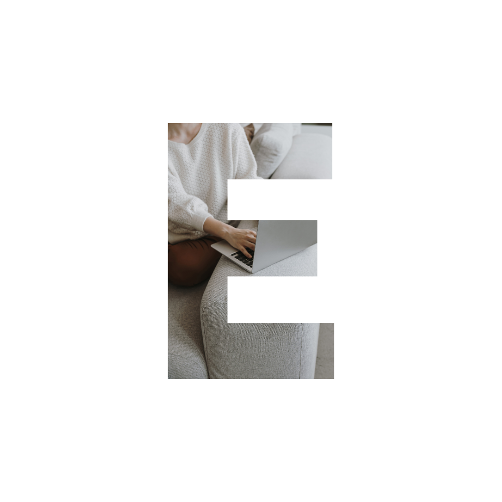 A person sitting on a sofa, using a silver laptop computer. They are wearing a cream sweater and brown pants.